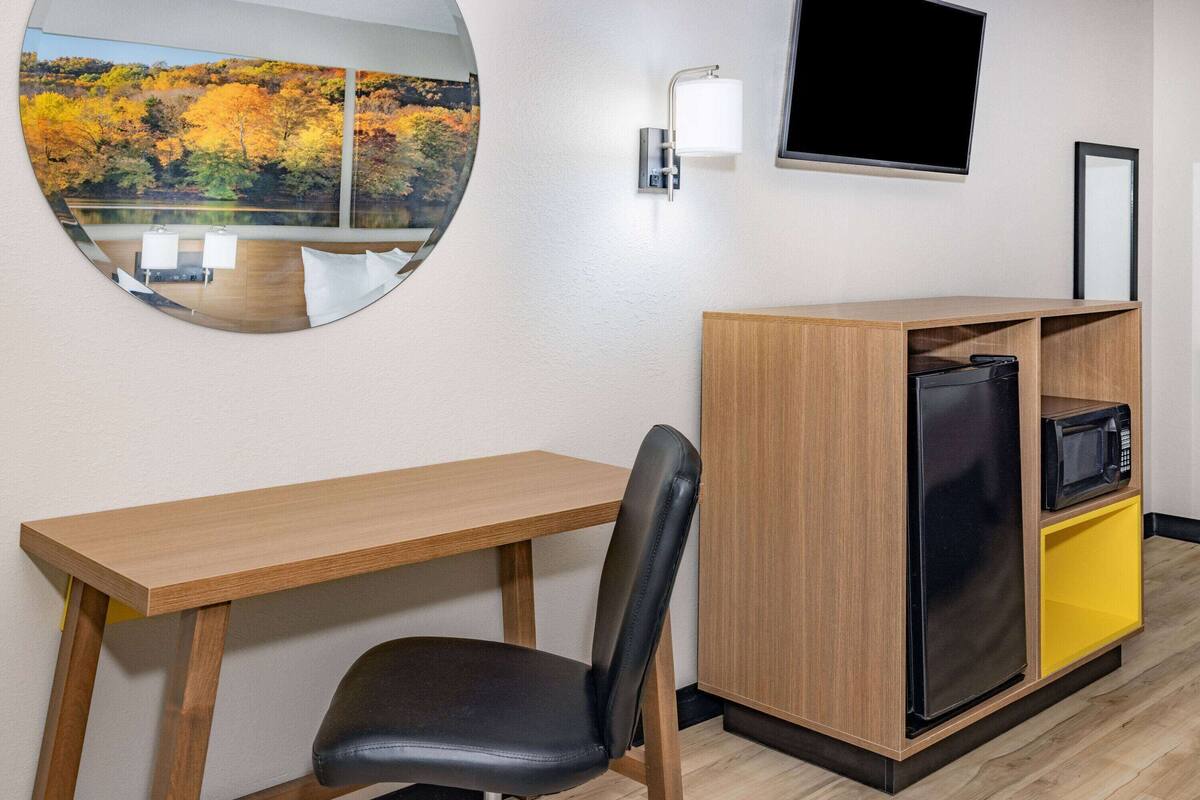 A hotel room with a wooden desk, a black chair, a small refrigerator, a microwave, and a TV on the wall.