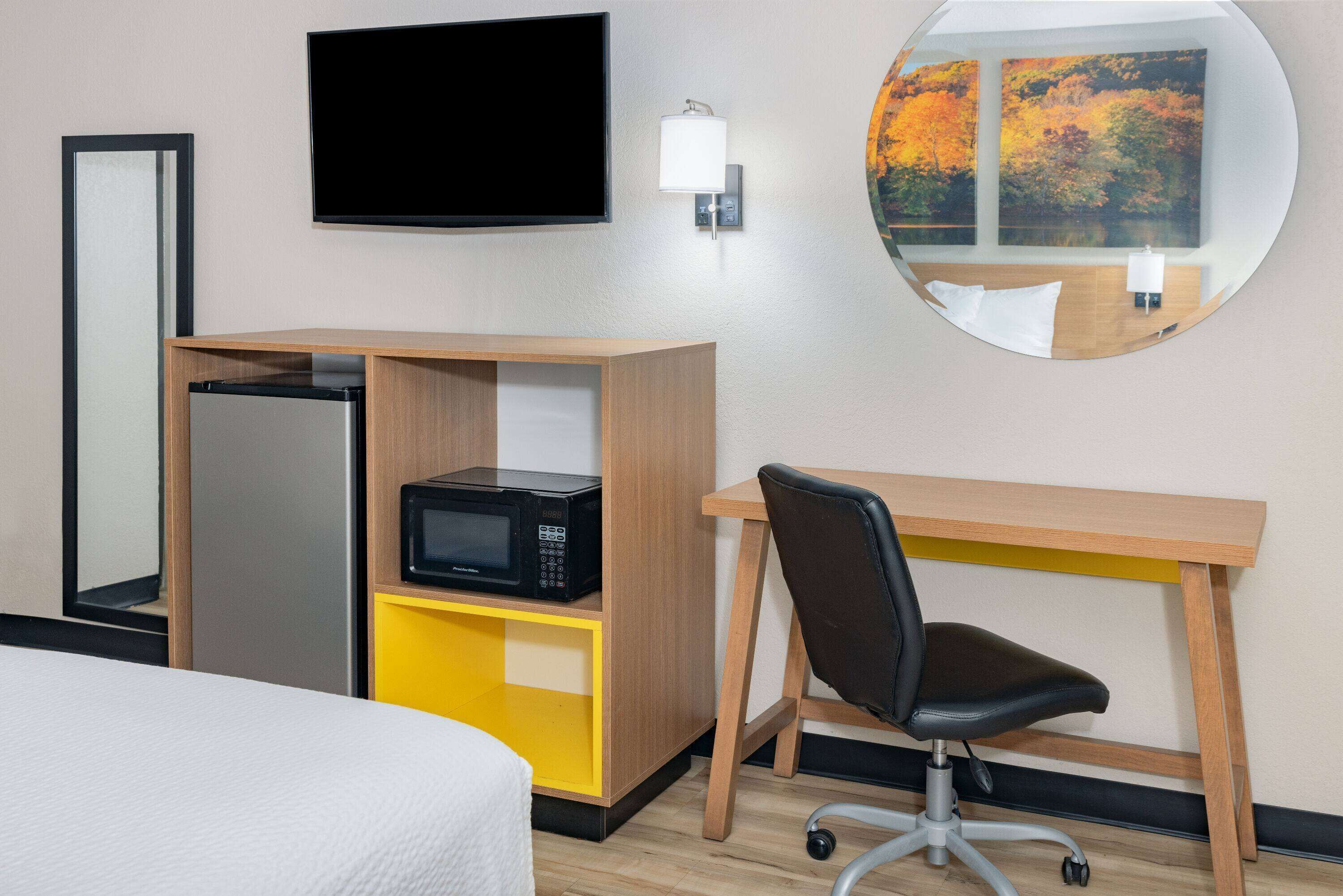 A hotel room with a bed, a desk with a chair, a microwave, a TV, a wall-mounted lamp, and a round mirror.
