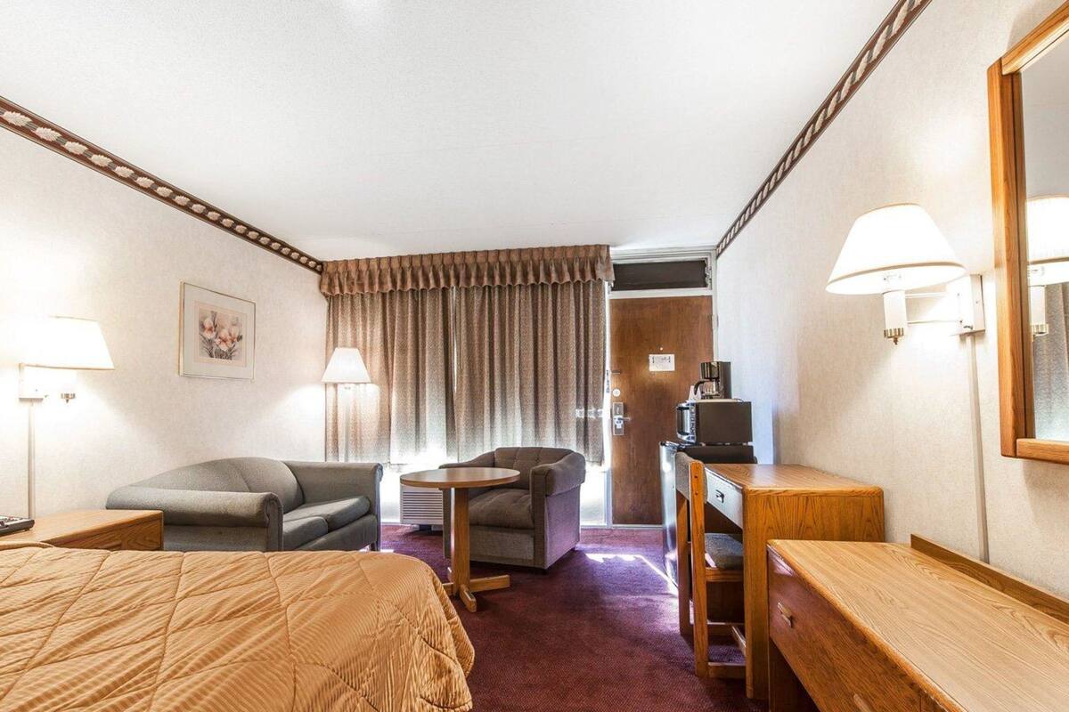 A hotel room with a bed, two armchairs, a small table, a desk, a mirror, and a window with curtains.