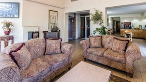 A spacious living area with two patterned sofas, a coffee table, and a TV. There is a kitchen area in the background with a dining table and chairs.