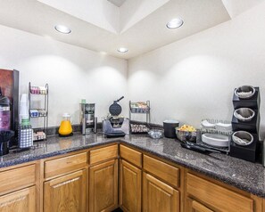 A hotel kitchen with granite countertops, wooden cabinets, a coffee machine, a waffle maker, and various food and drink dispensers.