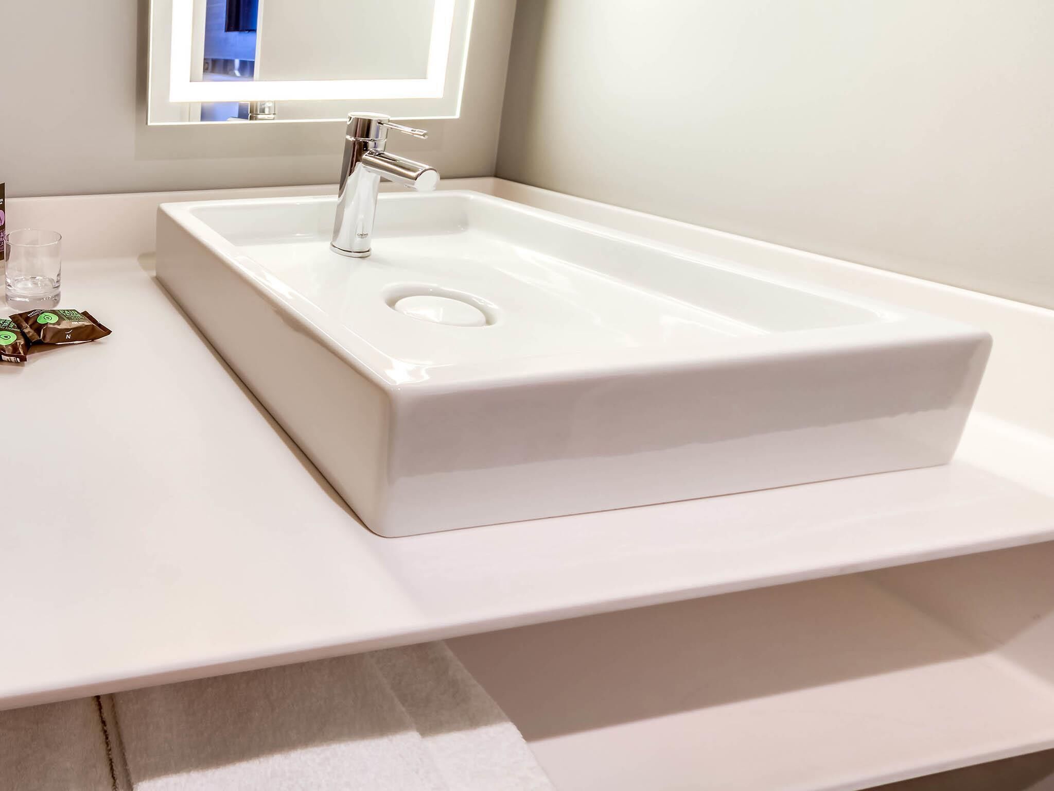 A white bathroom sink with a modern faucet, a mirror above it, and a small glass and a packet of toiletries on the counter.
