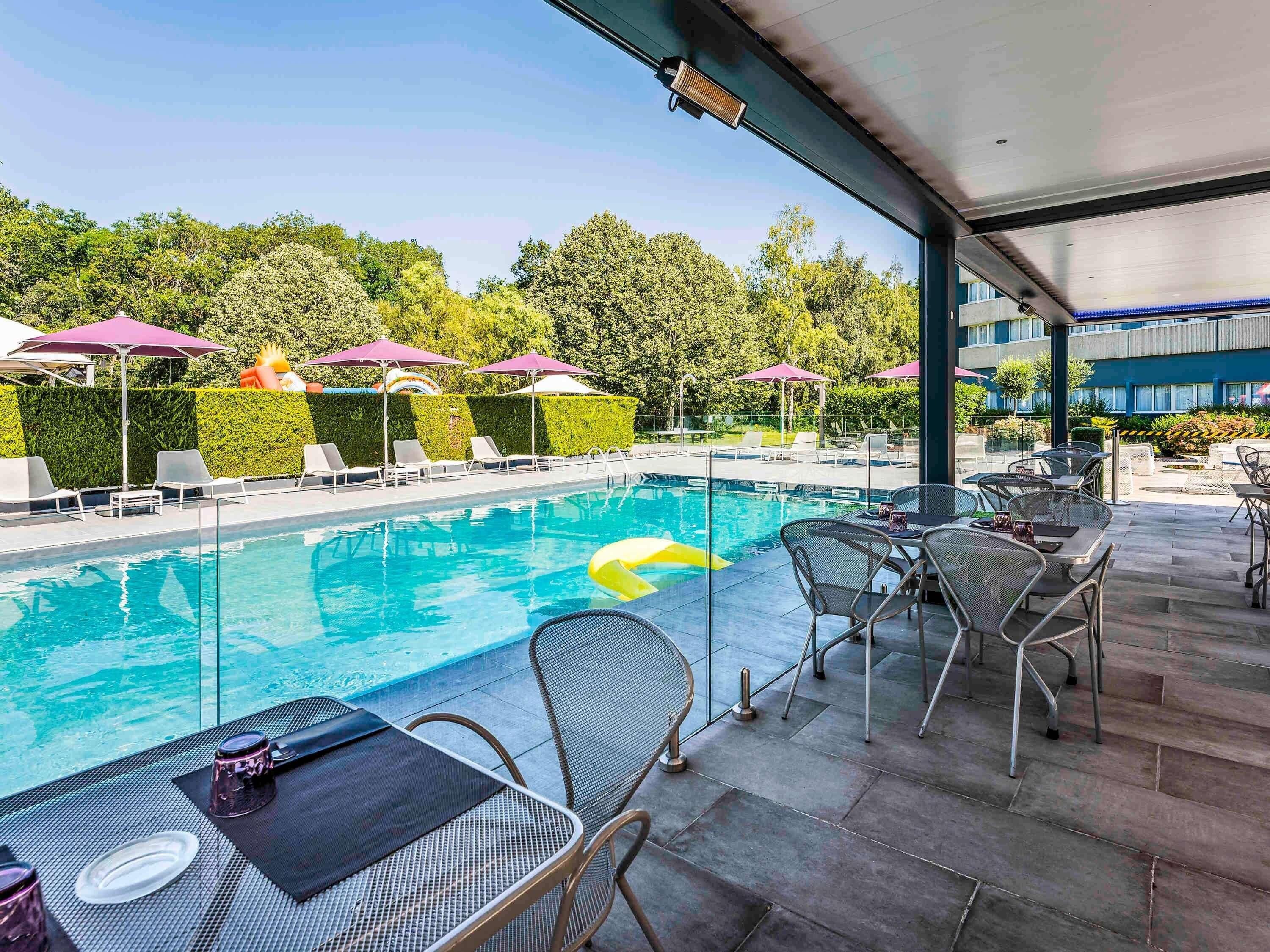 Zona de piscina con sillones de descanso y sombrillas, un área de comedor con mesas y sillas, y vista de un edificio al fondo.