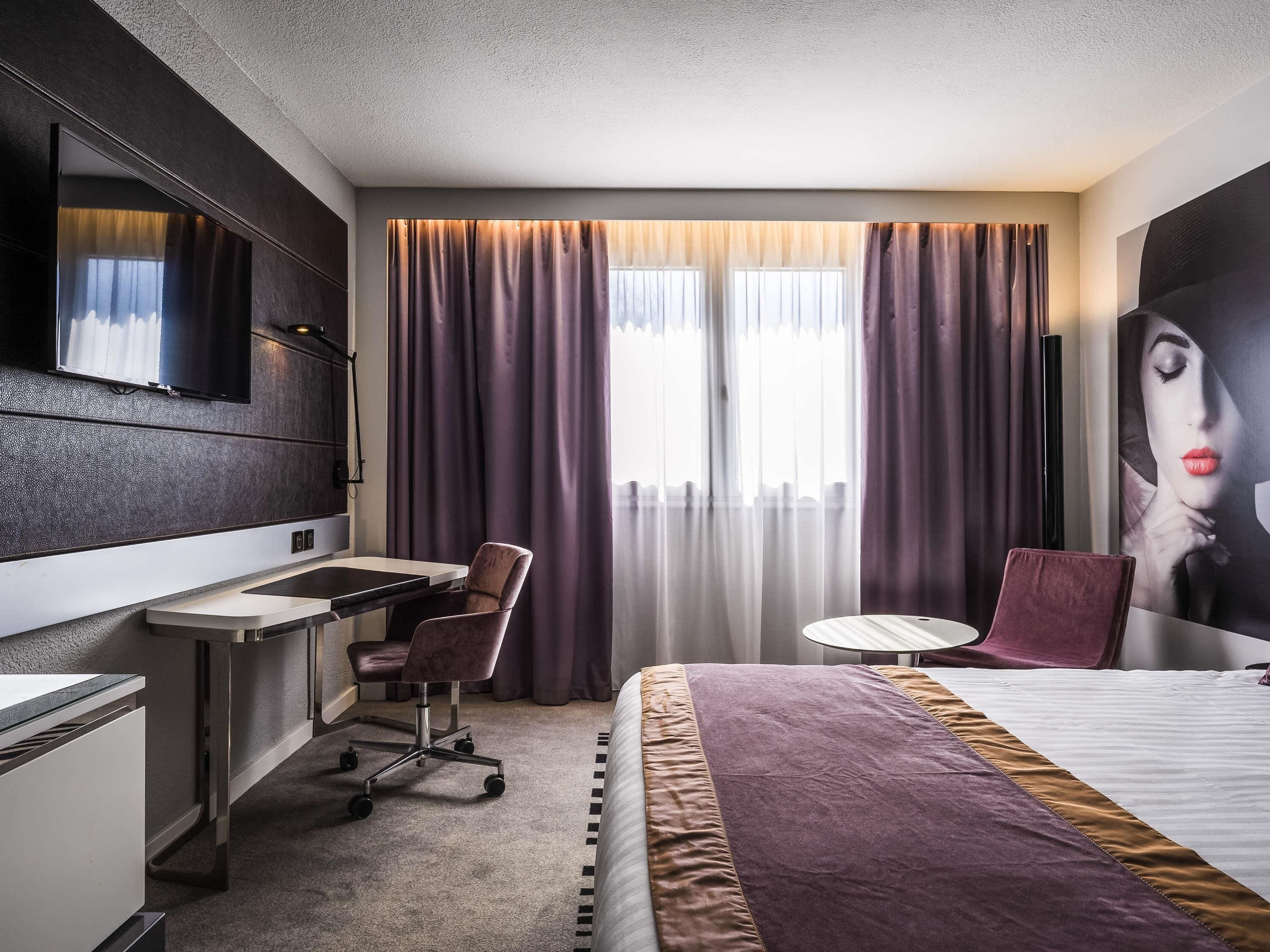 A hotel room with a desk, chair, bed, and a large window with curtains.