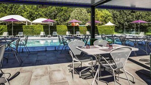 Zona de piscina exterior con sillas de descanso y sombrillas, rodeada de vegetación.