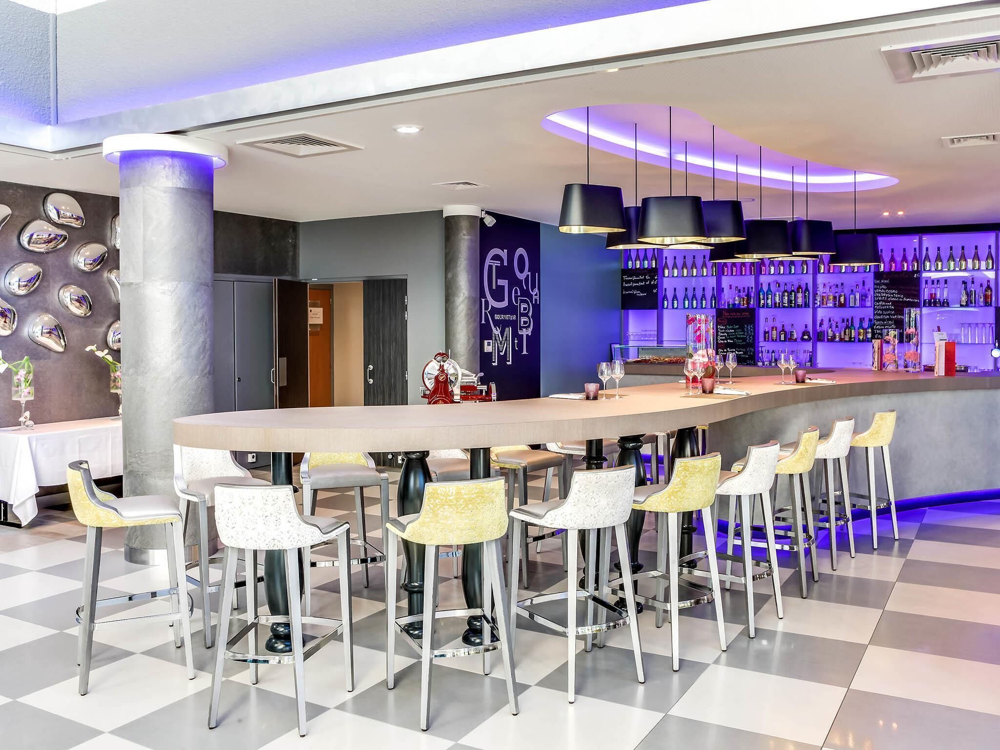 Una zona de bar con una barra larga, varios taburetes y una pared llena de botellas de vino.