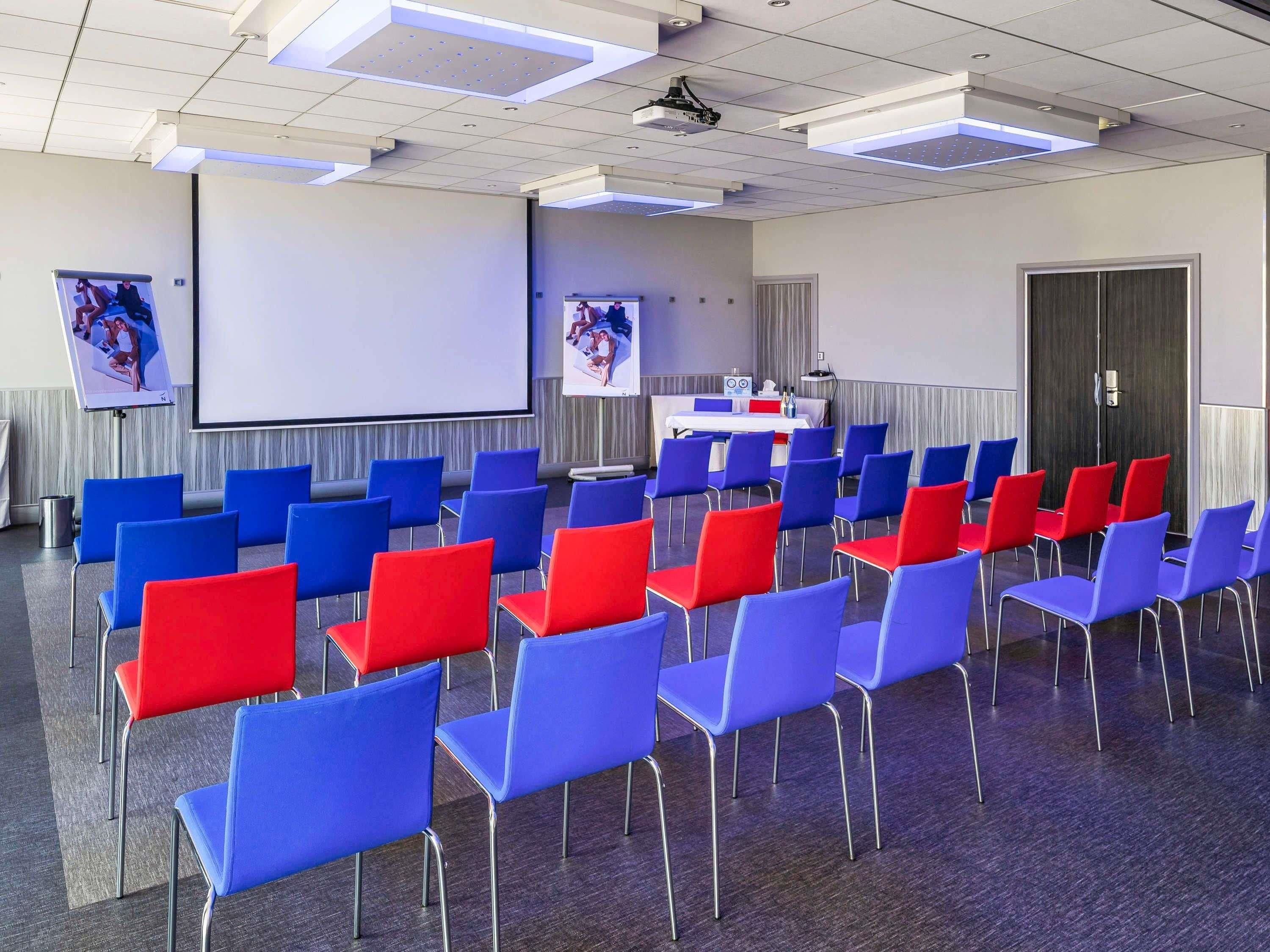 A conference room with rows of blue and red chairs, a projector screen, and a table with a white tablecloth.