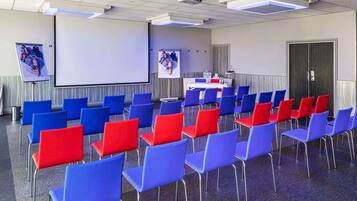 A conference room with rows of blue and red chairs, a projector screen, and a table with a white tablecloth.