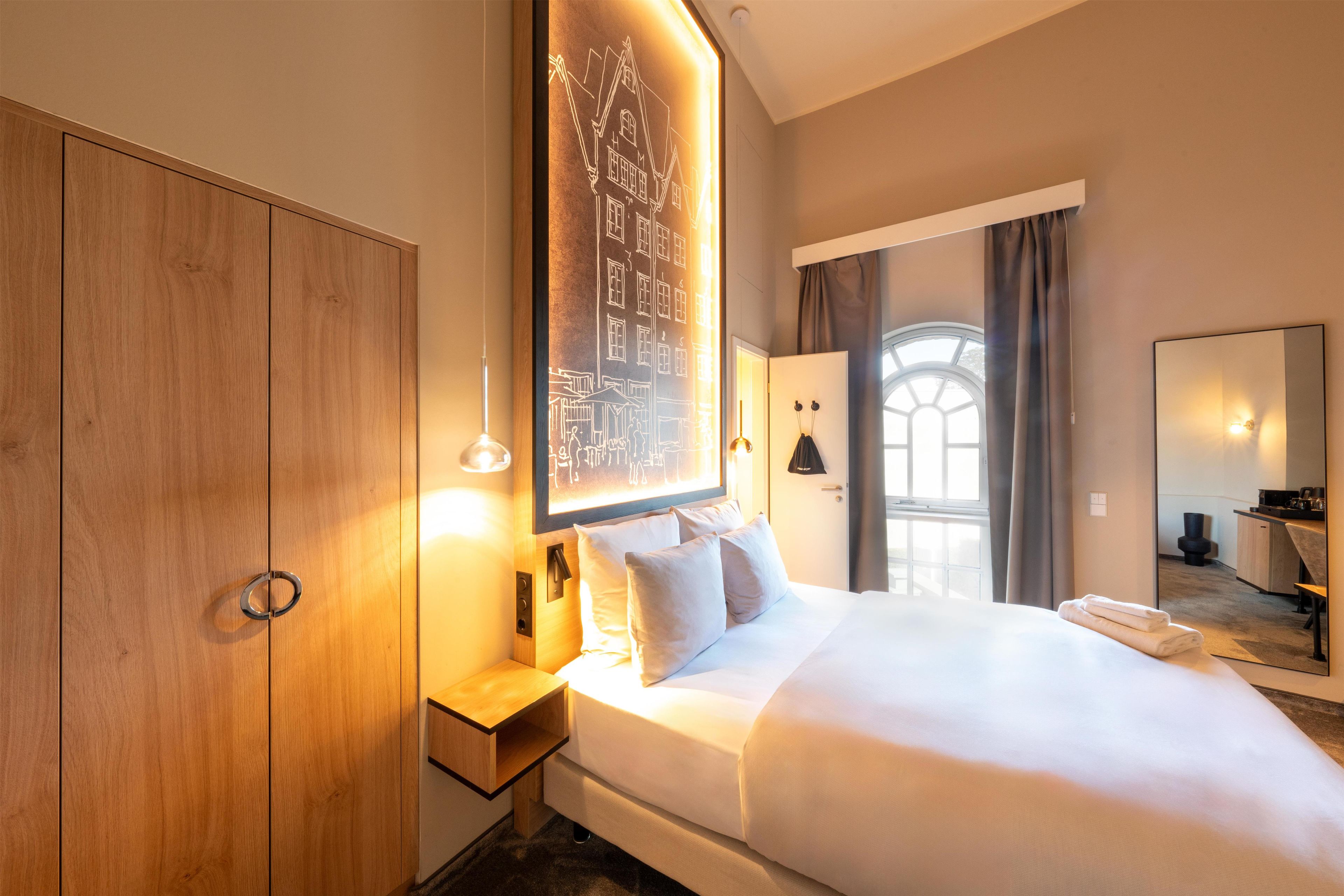 A hotel room with a large bed, a wooden wardrobe, a bedside table, a wall-mounted artwork, a window with curtains, and a bathroom visible through the door.