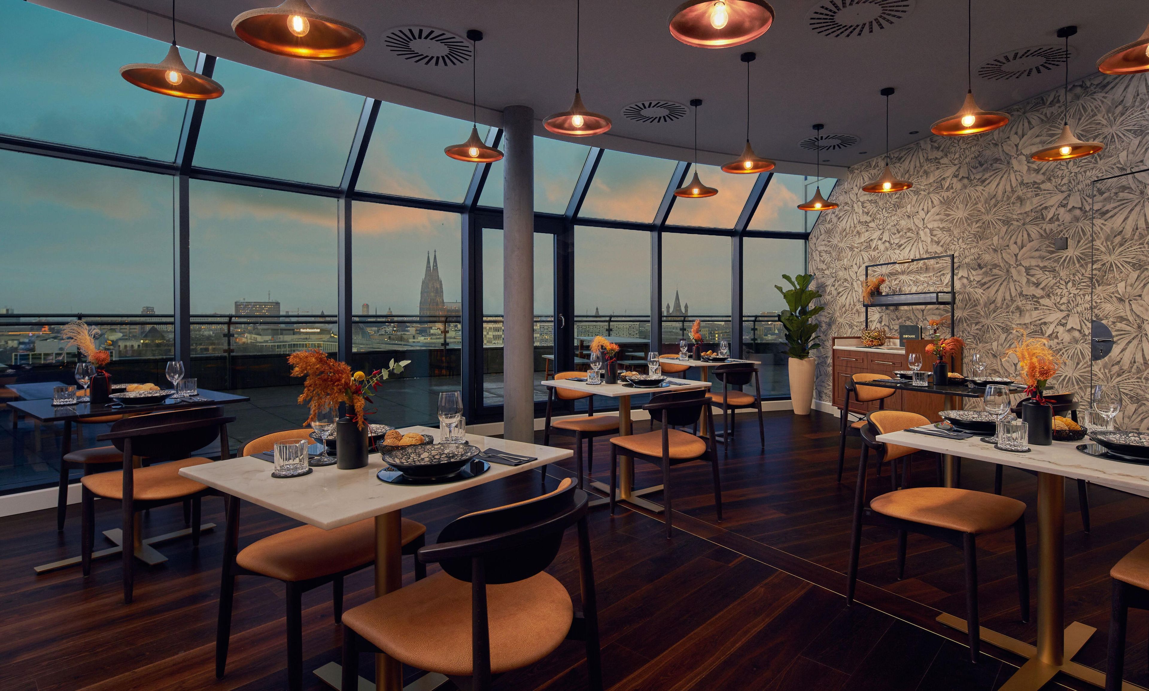 Ein Restaurant mit hohen Decken, großen Fenstern und Blick auf die Stadt. Die Tische sind mit Tellern, Gläsern und Blumen gedeckt.
