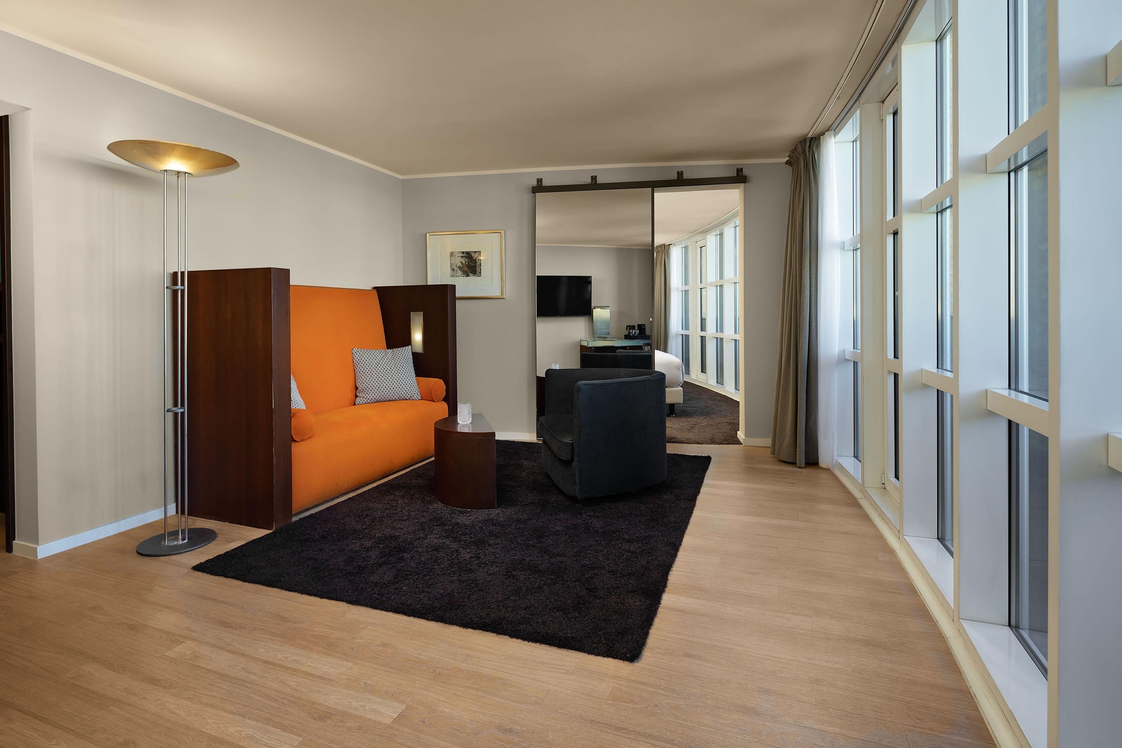 A modern hotel room with a wooden floor, an orange sofa, a black armchair, a small side table, a floor lamp, a flat-screen TV, and large windows with curtains.