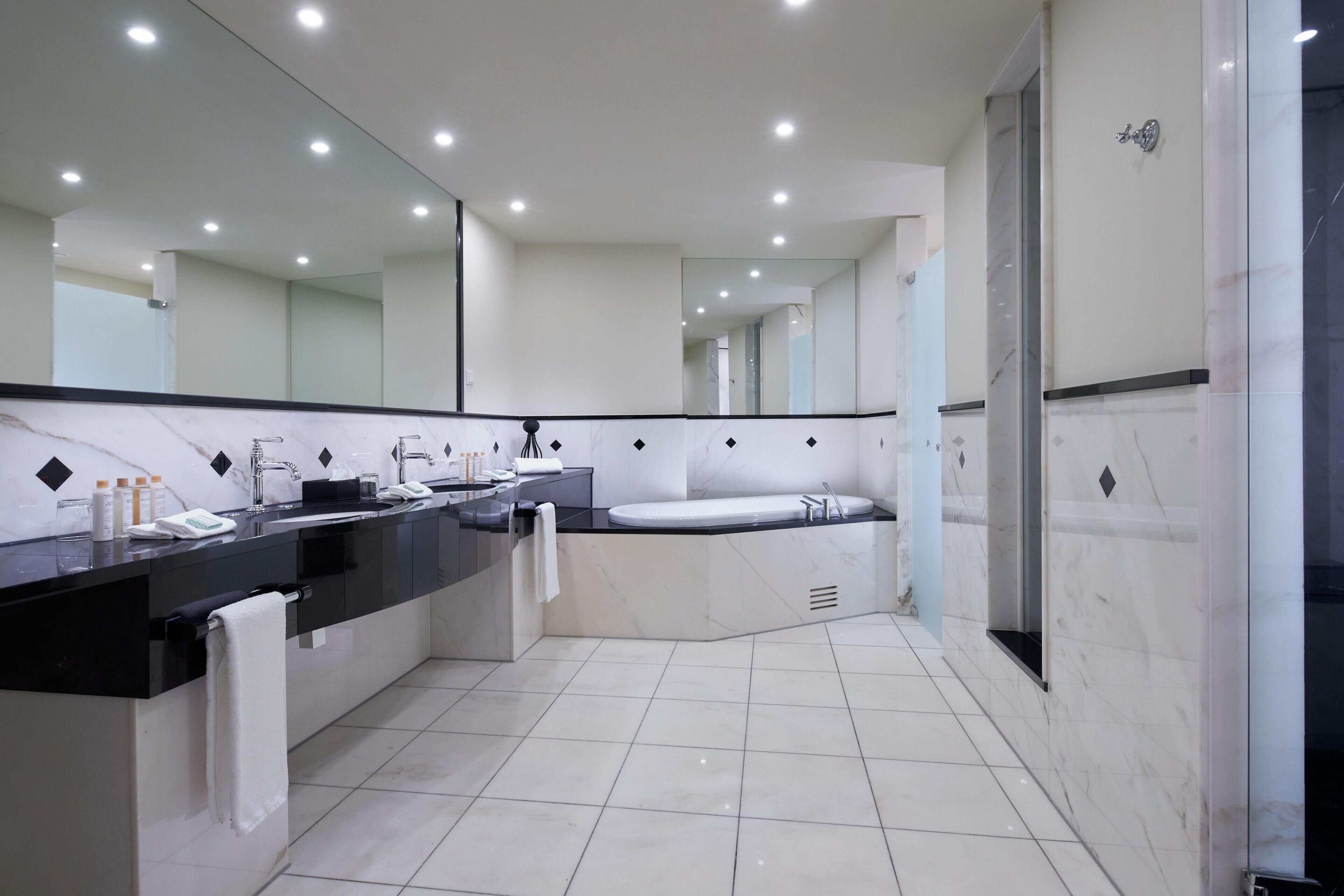 A modern bathroom with a large mirror, two sinks, a bathtub, and a shower area.