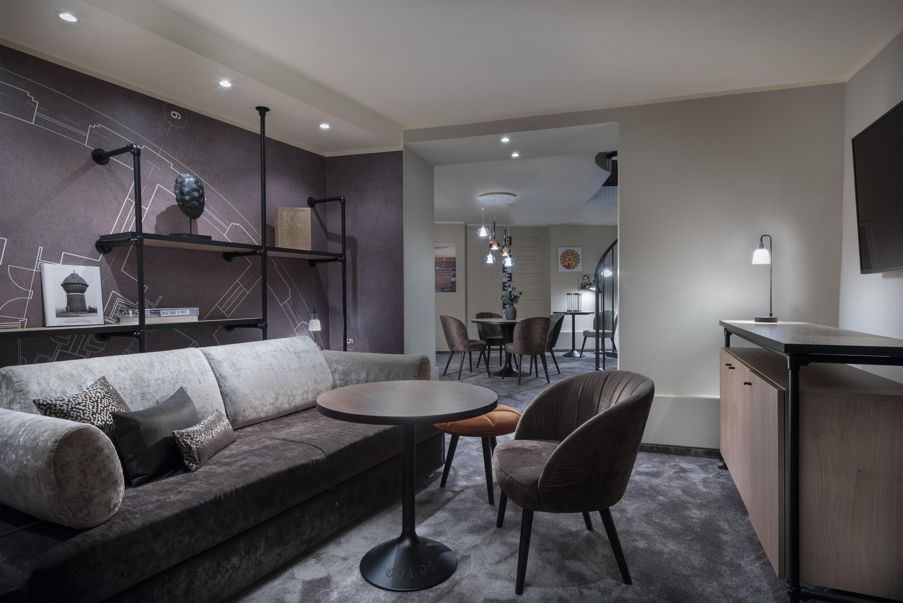 A modern living room with a sofa, round coffee table, and chairs. There is a bookshelf with decorative items and a dining area in the background.