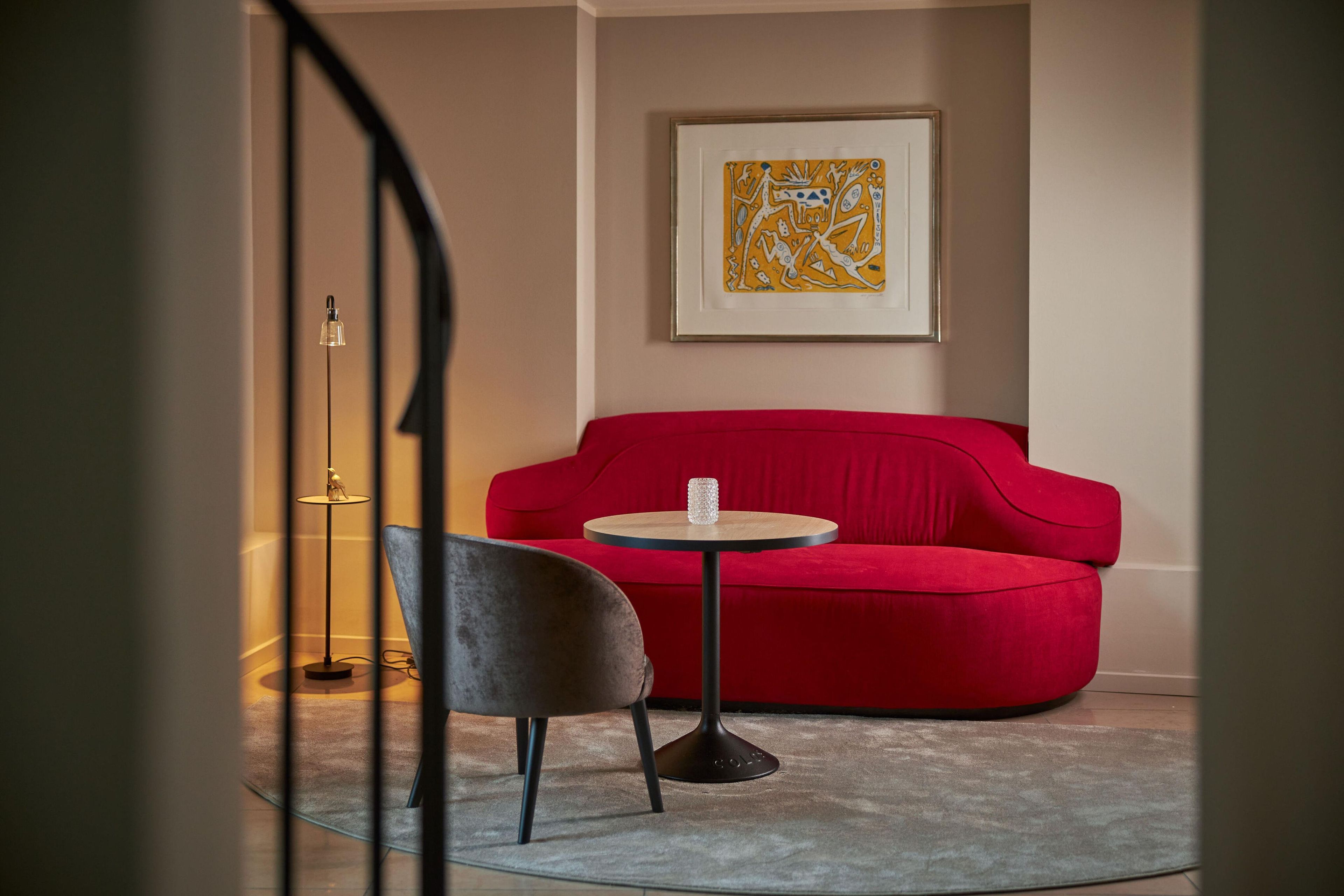 Un sofá rojo, una mesita redonda pequeña, una silla gris y una lámpara de pie en una habitación con un cuadro abstracto enmarcado en la pared.