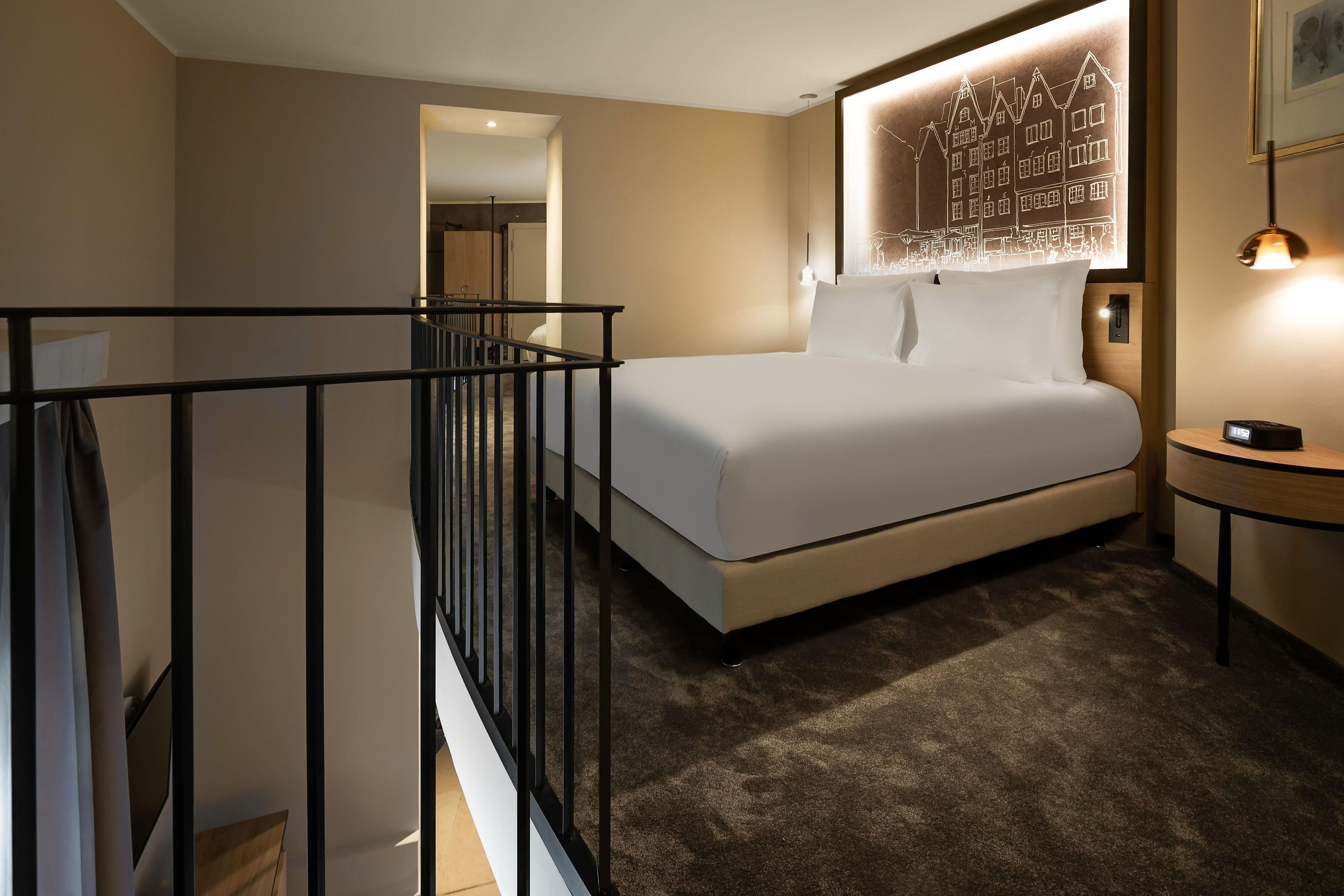 A hotel room with a bed, a bedside table, a nightstand, a wall-mounted light, and a ceiling light.