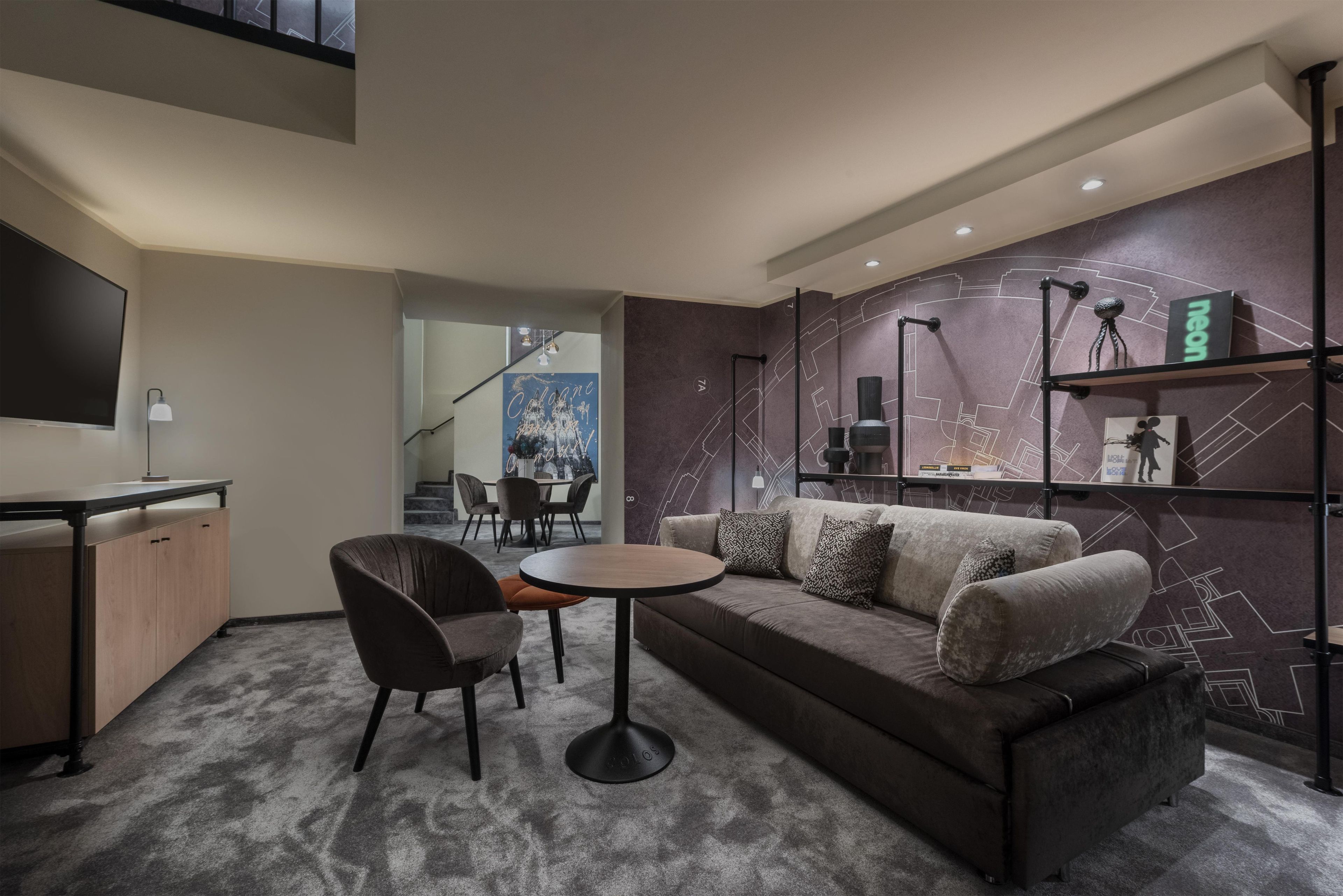 Un salon moderne avec un canapé, une table basse et des fauteuils. Un téléviseur à écran plat est fixé au mur, un escalier mène à un autre niveau, et un mur décoratif présente des motifs géométriques.