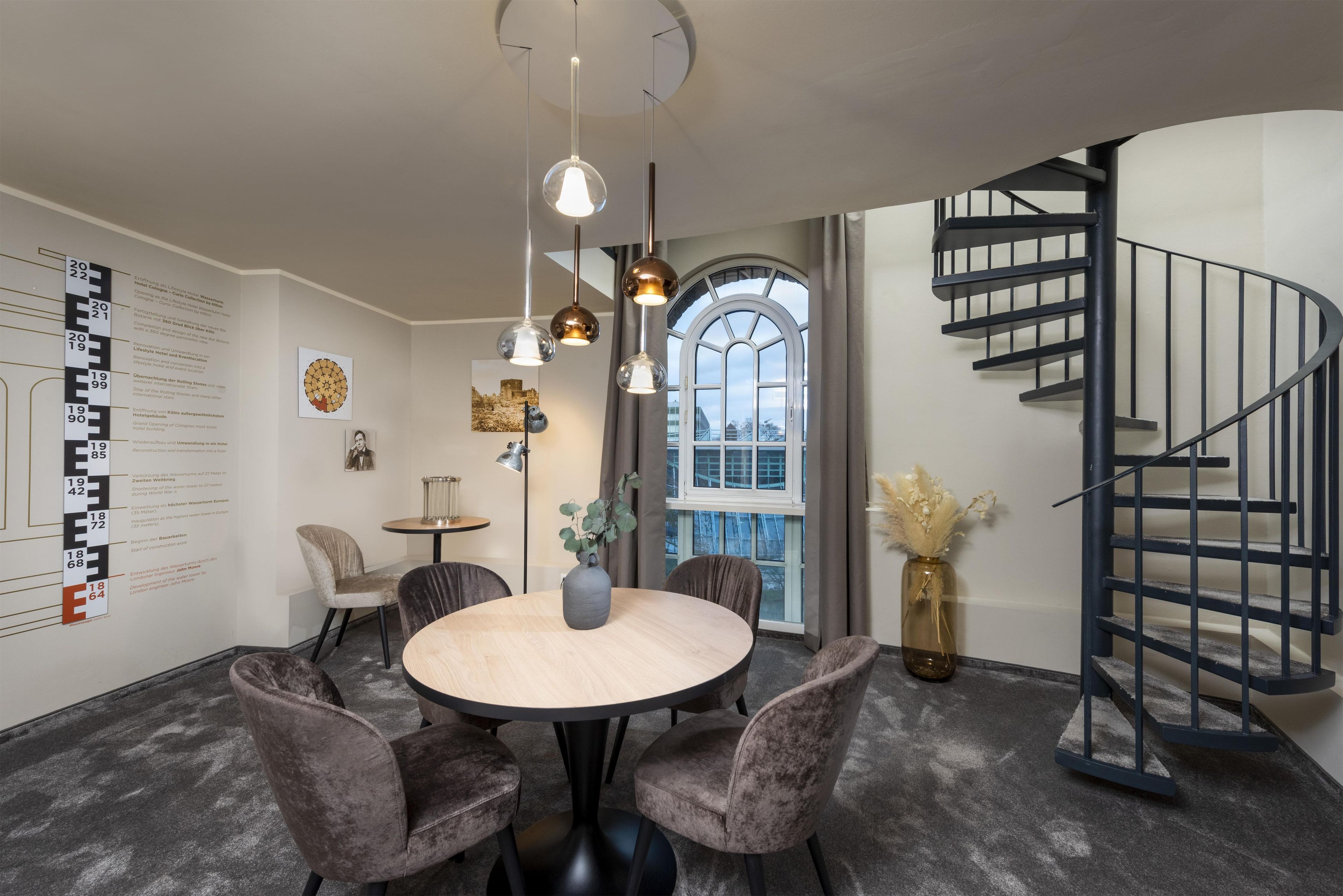 A modern dining area with a round table, grey upholstered chairs, pendant lighting, and a spiral staircase.