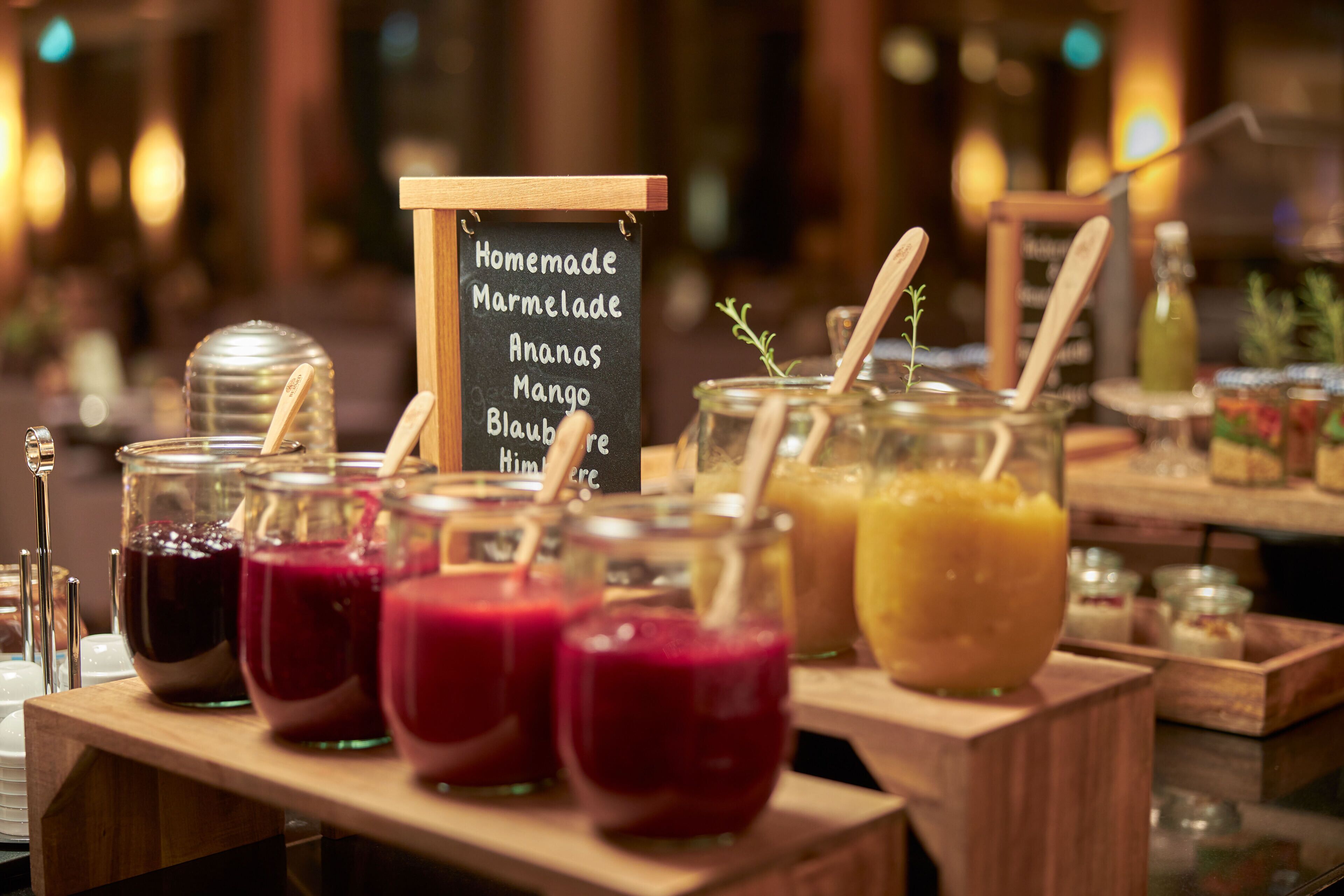 A buffet with various homemade jams and jellies, including mango and blueberry, displayed in glass jars with wooden spoons.