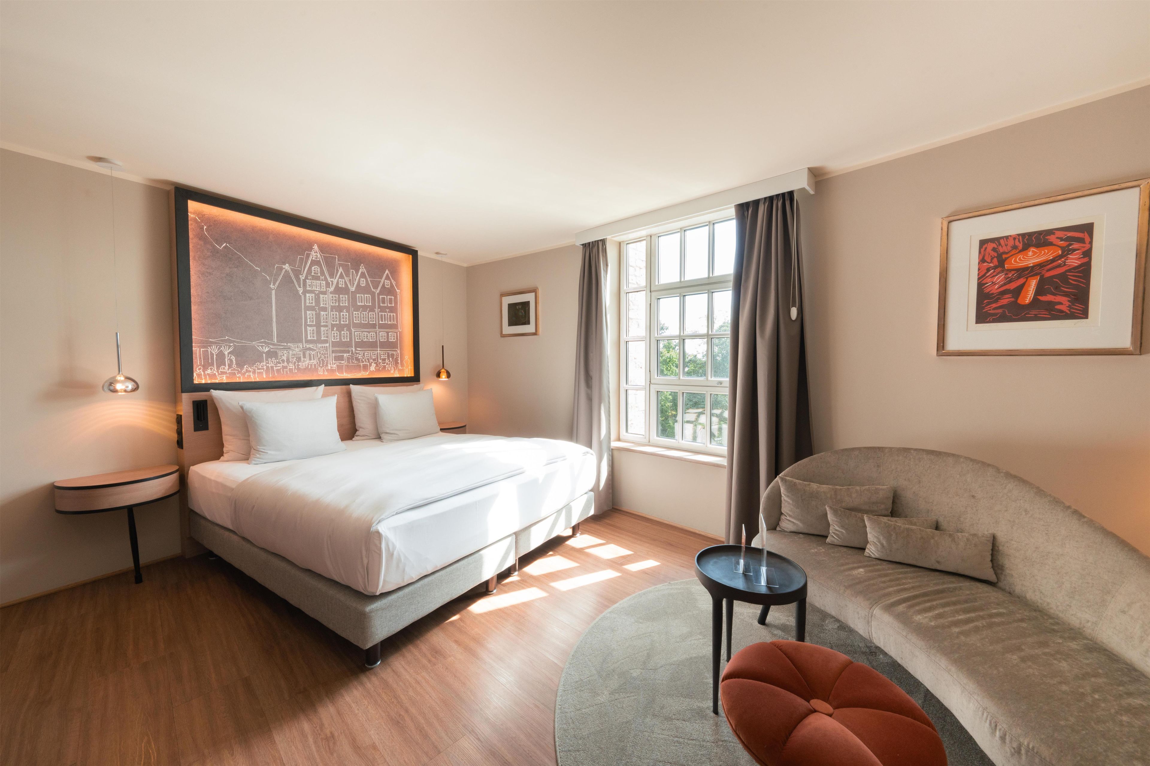 A hotel room with a bed, a sofa, a small table, a painting, and a window with curtains.