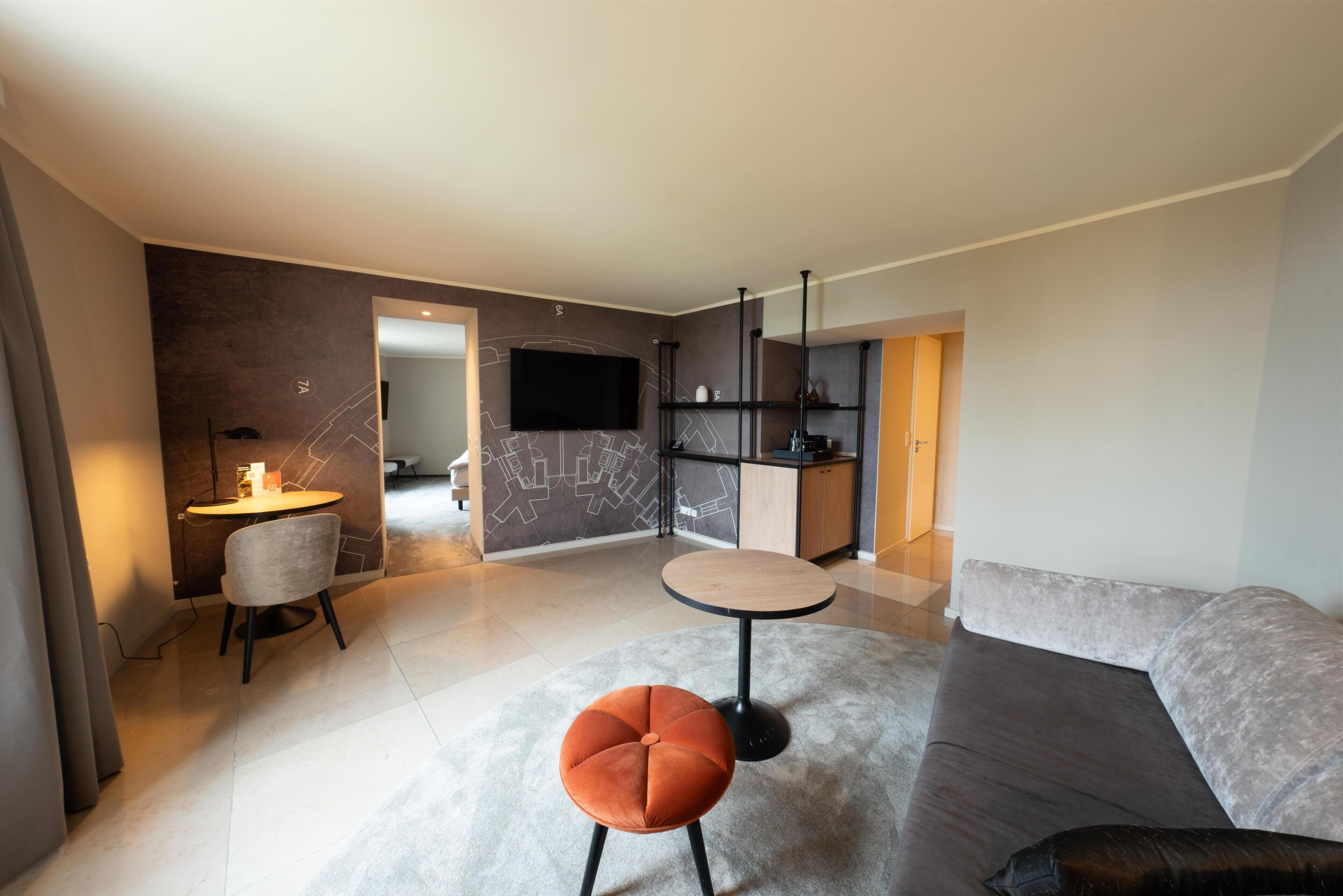 Een moderne woonkamer met een grijze bank, een rond poefje, een kleine tafel en een aan de muur bevestigde televisie.