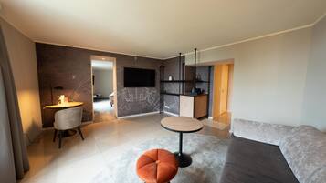 Een moderne woonkamer met een grijze bank, een rond poefje, een kleine tafel en een aan de muur bevestigde televisie.