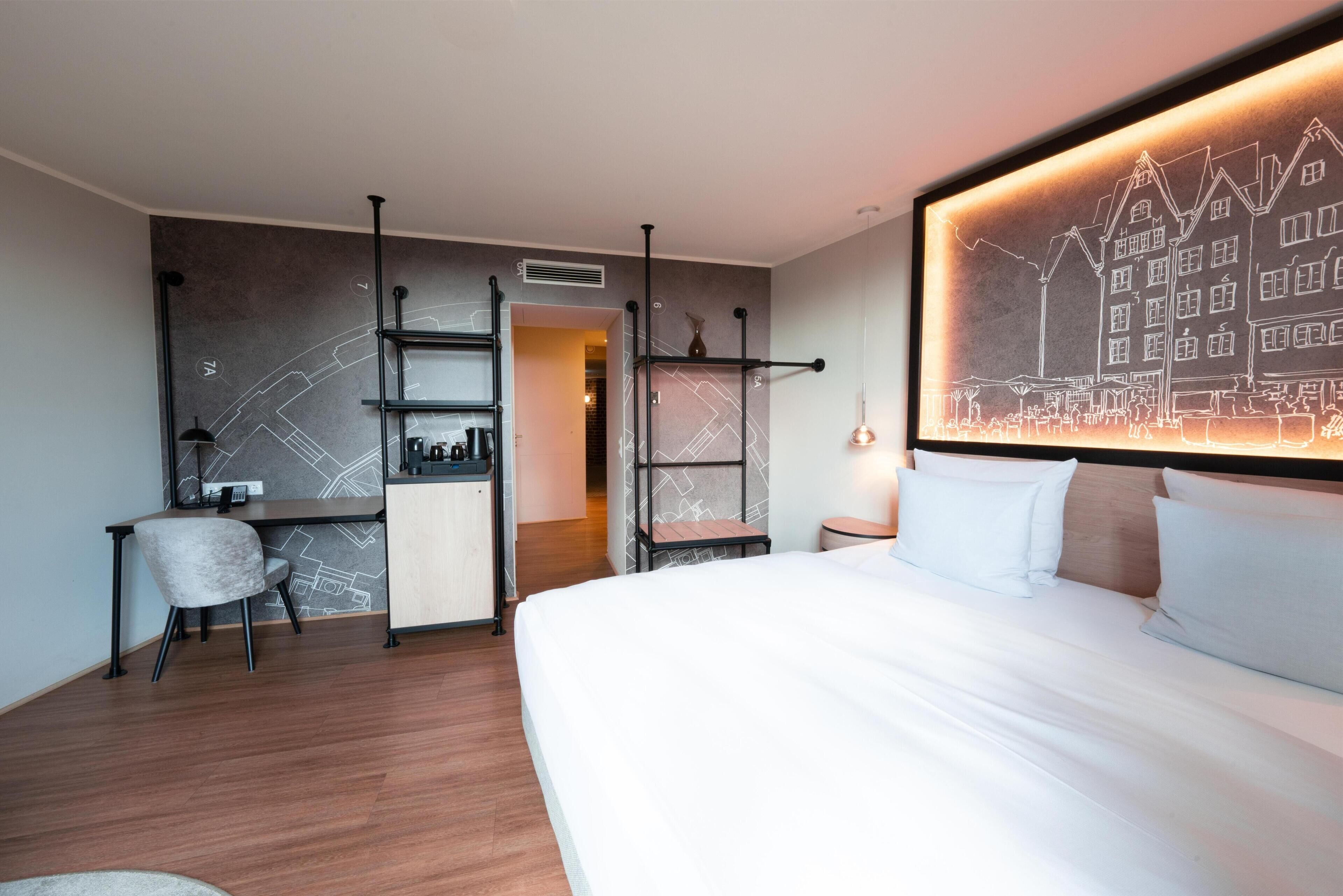A hotel room with a bed, a desk, a chair, a shelf, and a wall art piece.
