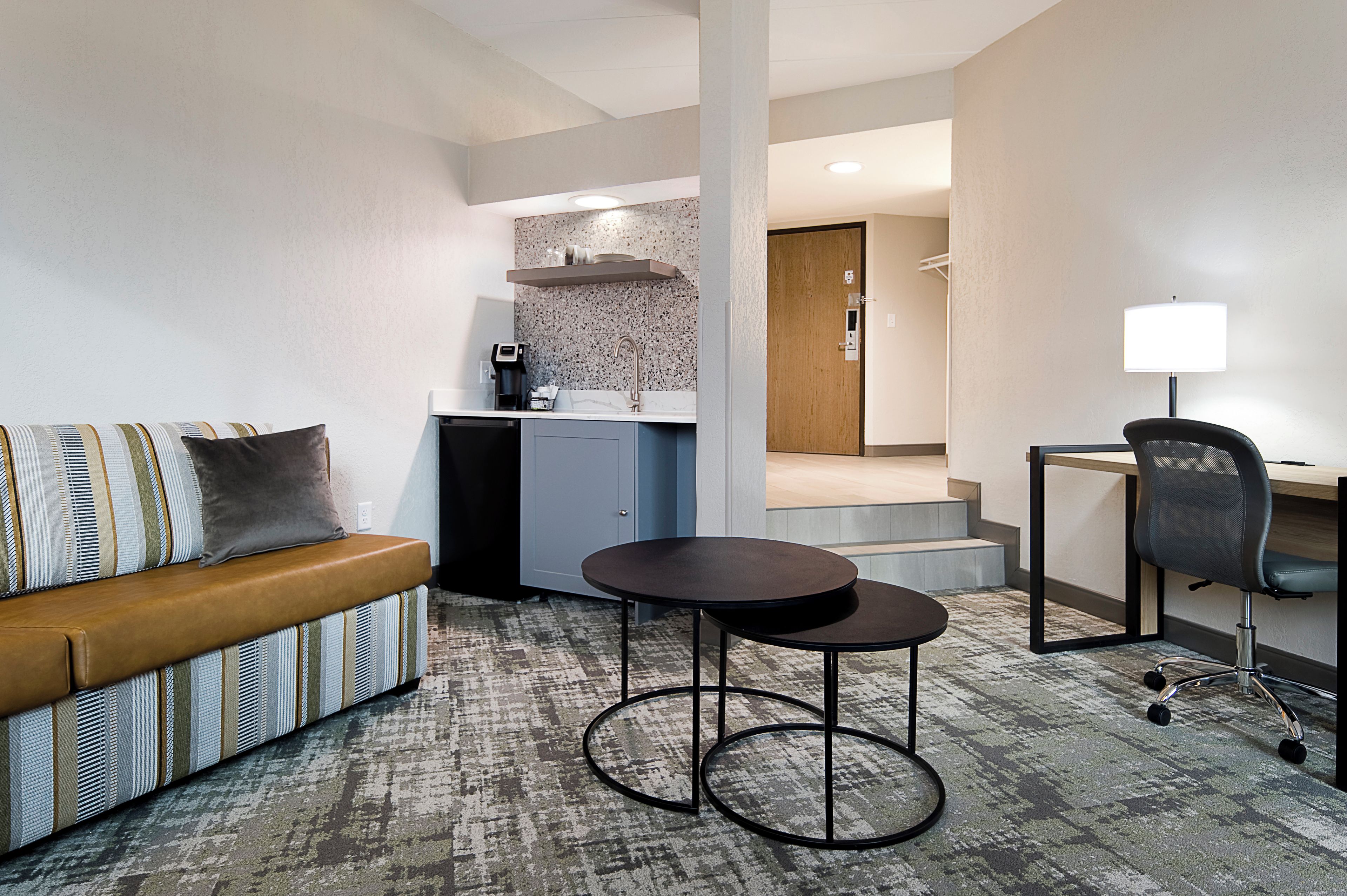 Una camera d'albergo con un divano, un piccolo tavolo rotondo, una scrivania con una sedia, una kitchenette e un bagno sullo sfondo.