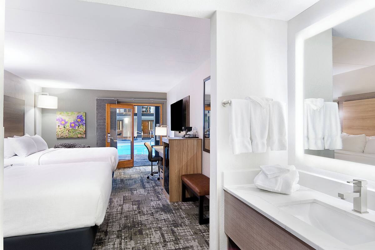 Ein Hotelzimmer mit zwei Betten, einem Schreibtisch, einem Stuhl, einem großen Spiegel, einem Badezimmer mit Waschbecken und Handtuchhalter sowie Blick auf einen Poolbereich.