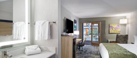 Habitación de hotel con un espejo grande, un lavamanos, un escritorio con una silla, una cama y un cuadro en la pared.