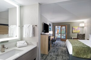 A hotel room with a large mirror, a sink, a desk with a chair, a bed, and a painting on the wall.