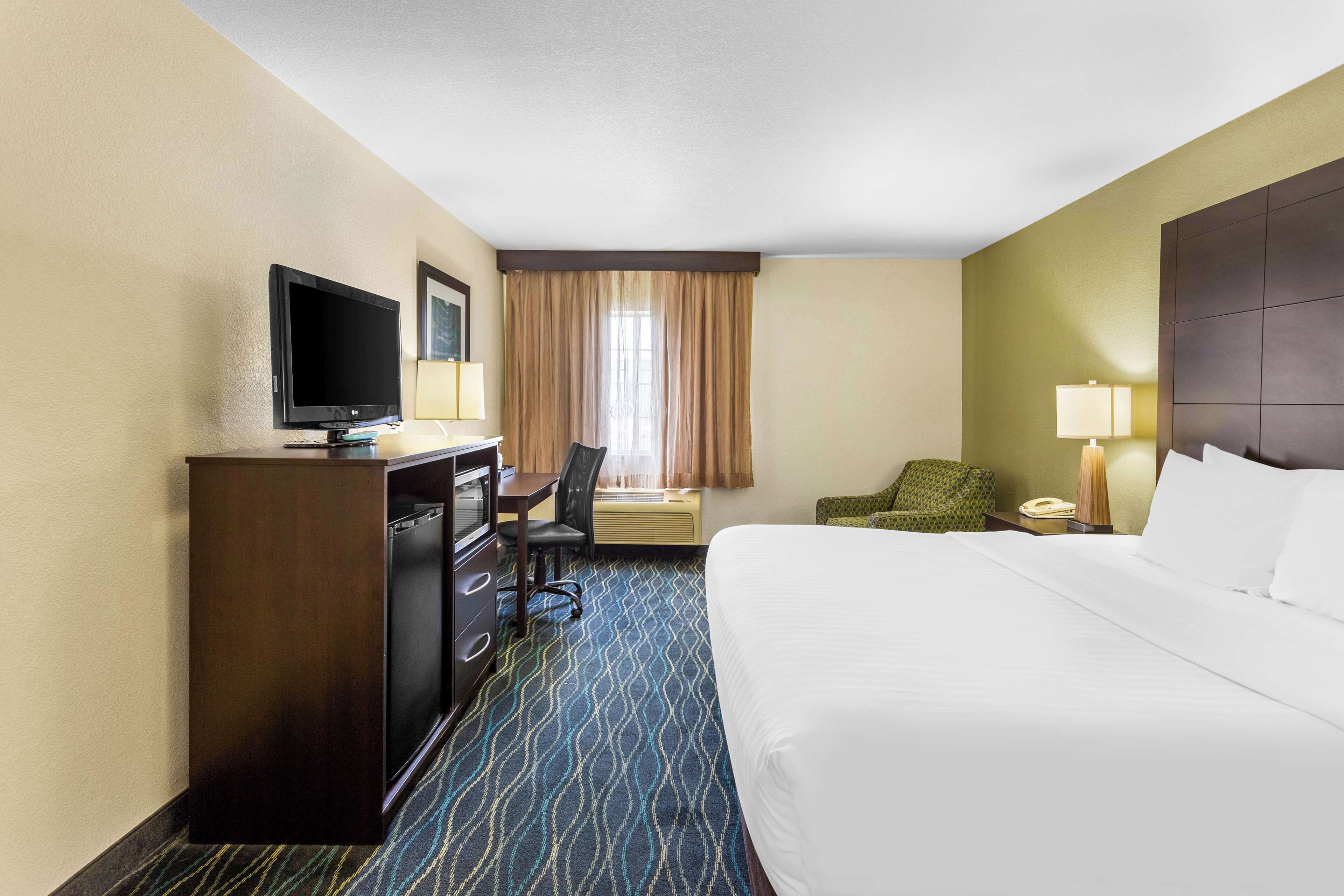 A hotel room with a bed, a desk with a television, a chair, a lamp, and a window with curtains.