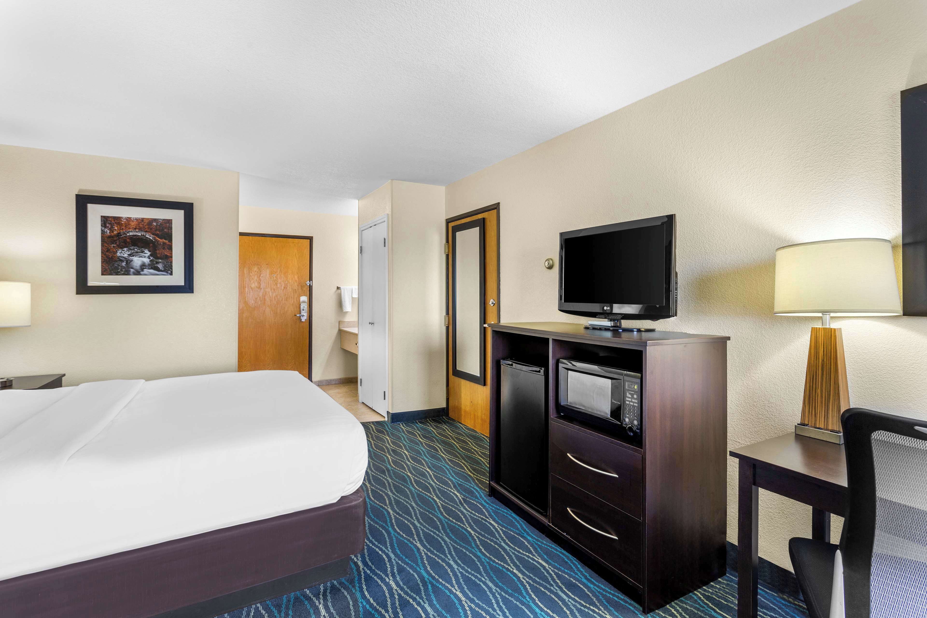 A hotel room with a bed, a television, a desk with a lamp, and a minibar.