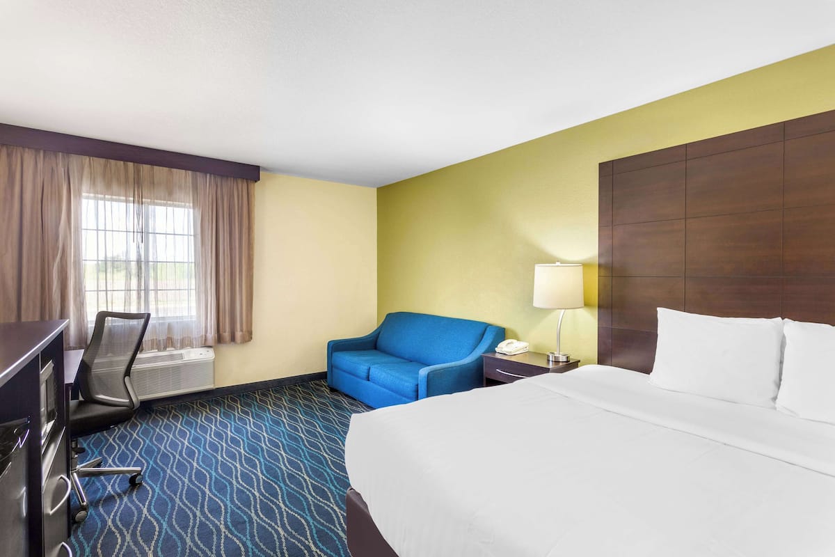 A hotel room with a large bed, a desk with a chair, a blue sofa, a lamp, and a window with curtains.