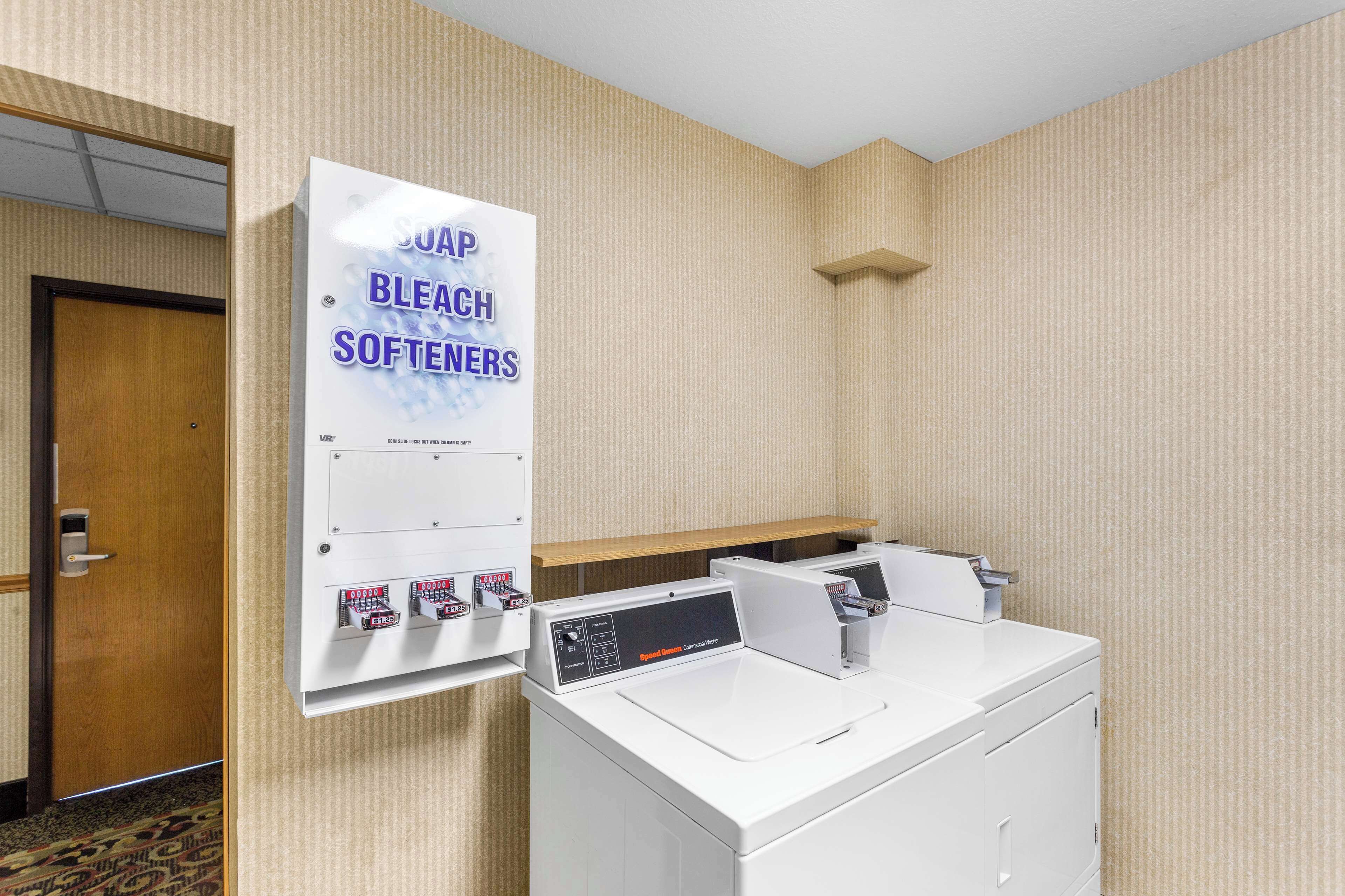 A hotel room with a wall-mounted washer and dryer, a sign advertising 'SUDAP BLEACH SOFTENERS', and a wooden door.