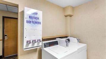 A hotel room with a wall-mounted washer and dryer, a sign advertising 'SUDAP BLEACH SOFTENERS', and a wooden door.