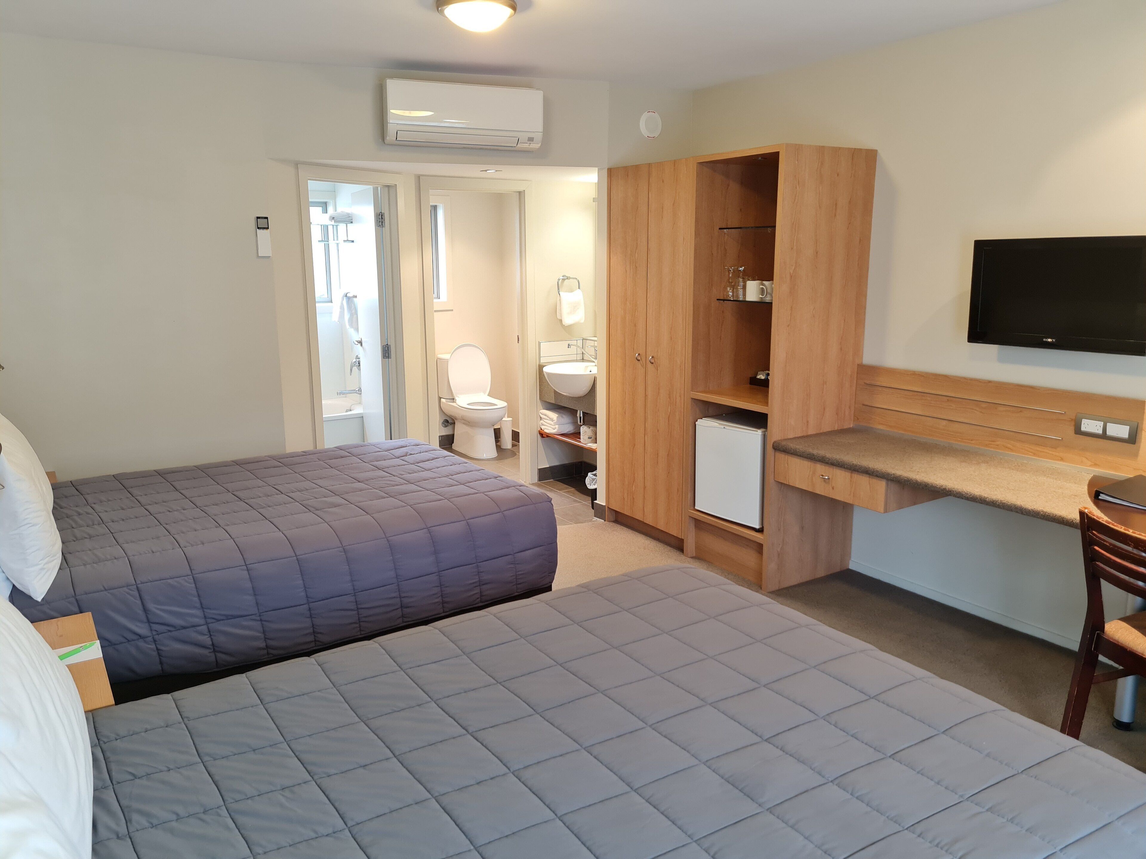 A bedroom with two beds, a wooden wardrobe, a small kitchenette, and a TV mounted on the wall.