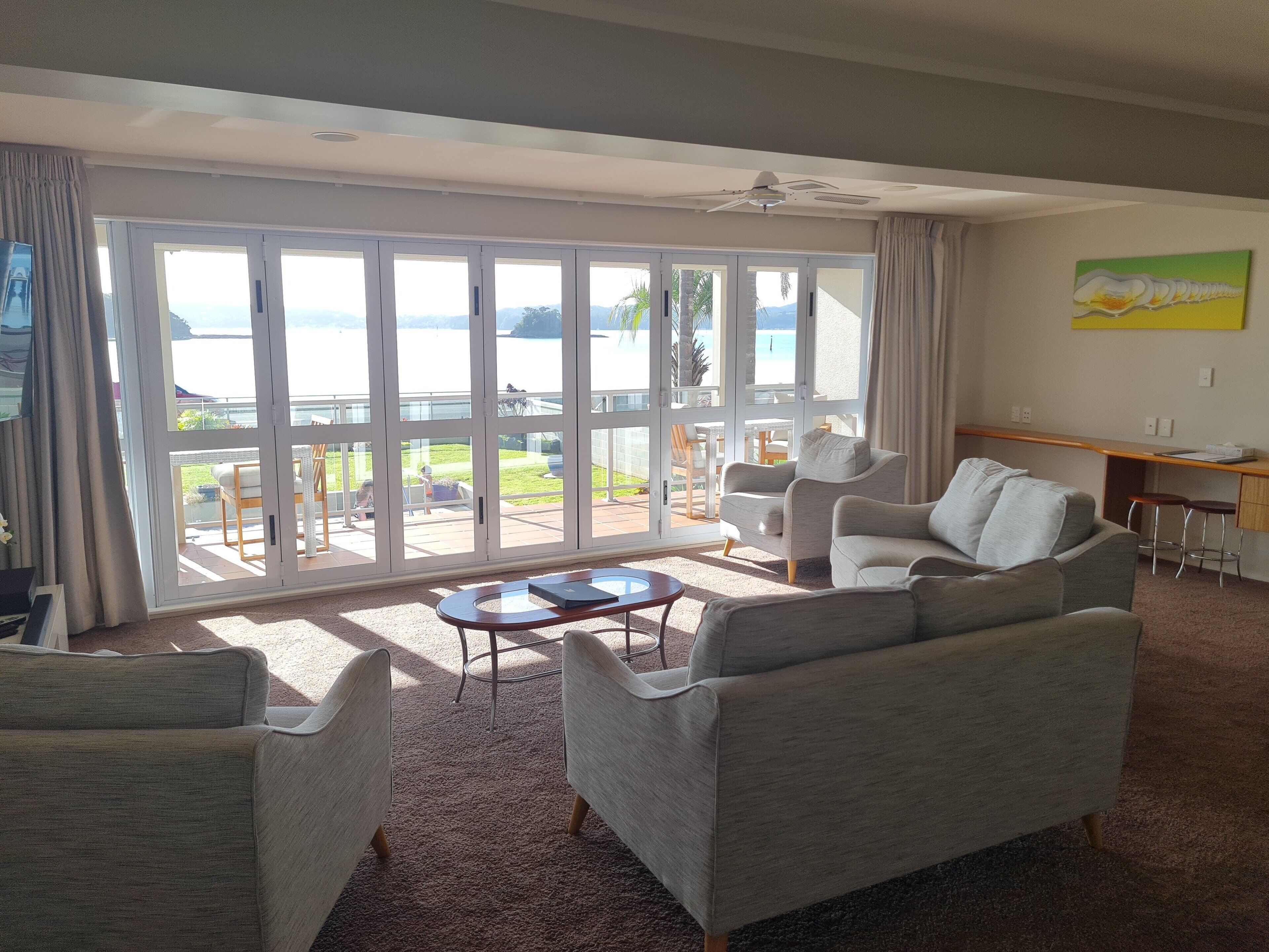 A living room with a large sliding glass door leading to a balcony with a view of the sea. There are two armchairs, a coffee table, and a desk with a chair.