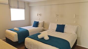 Habitación de hotel con dos camas, cada una con edredón azul y blanco, una mesita de noche pequeña y una ventana con persiana de color claro.