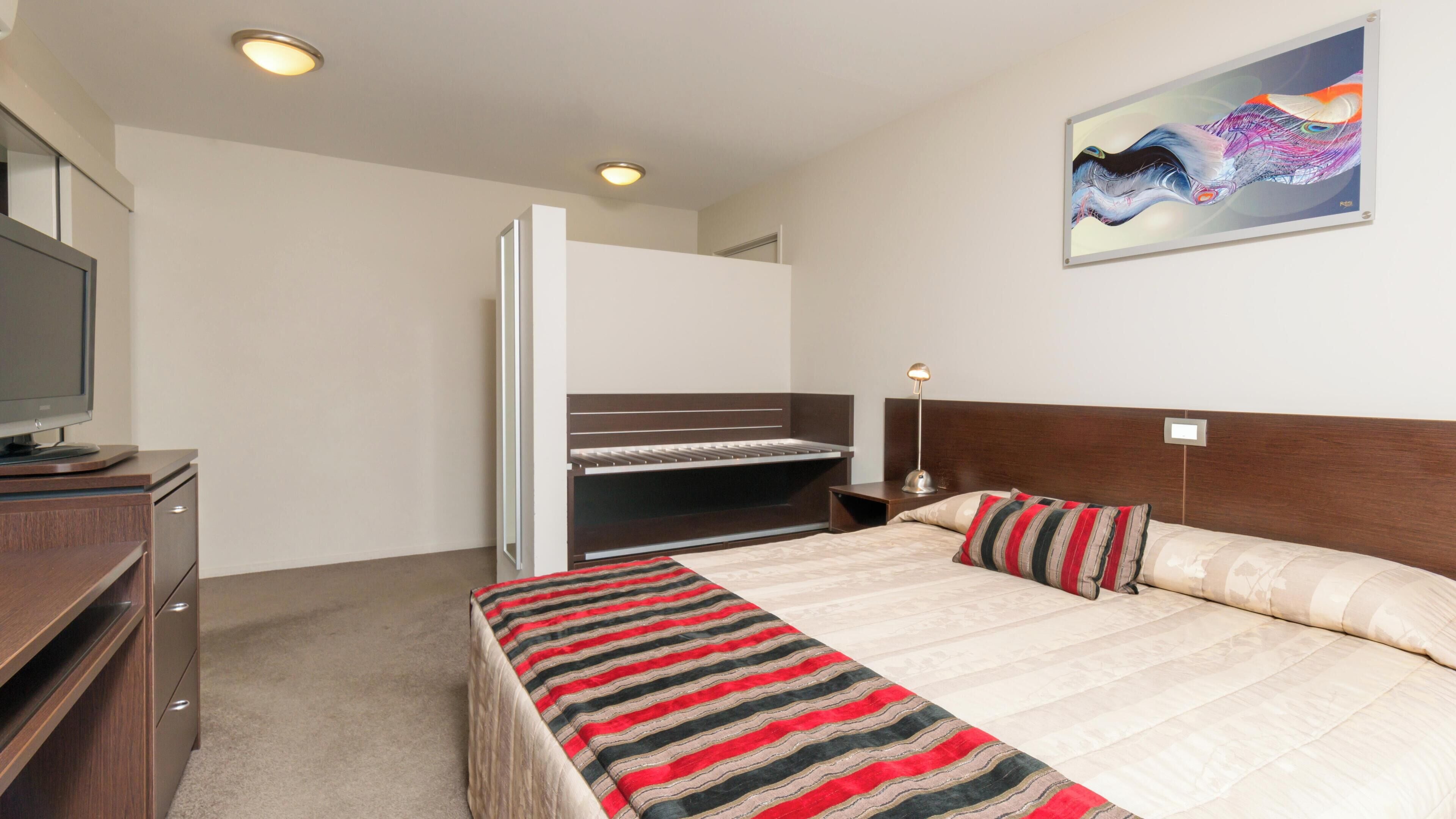 A hotel room with a bed, a desk, a television, and a painting on the wall.