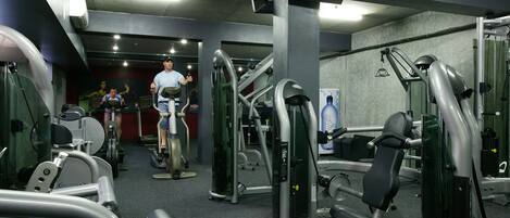 A gym with various exercise machines, a television mounted on the wall, and a person using a treadmill.