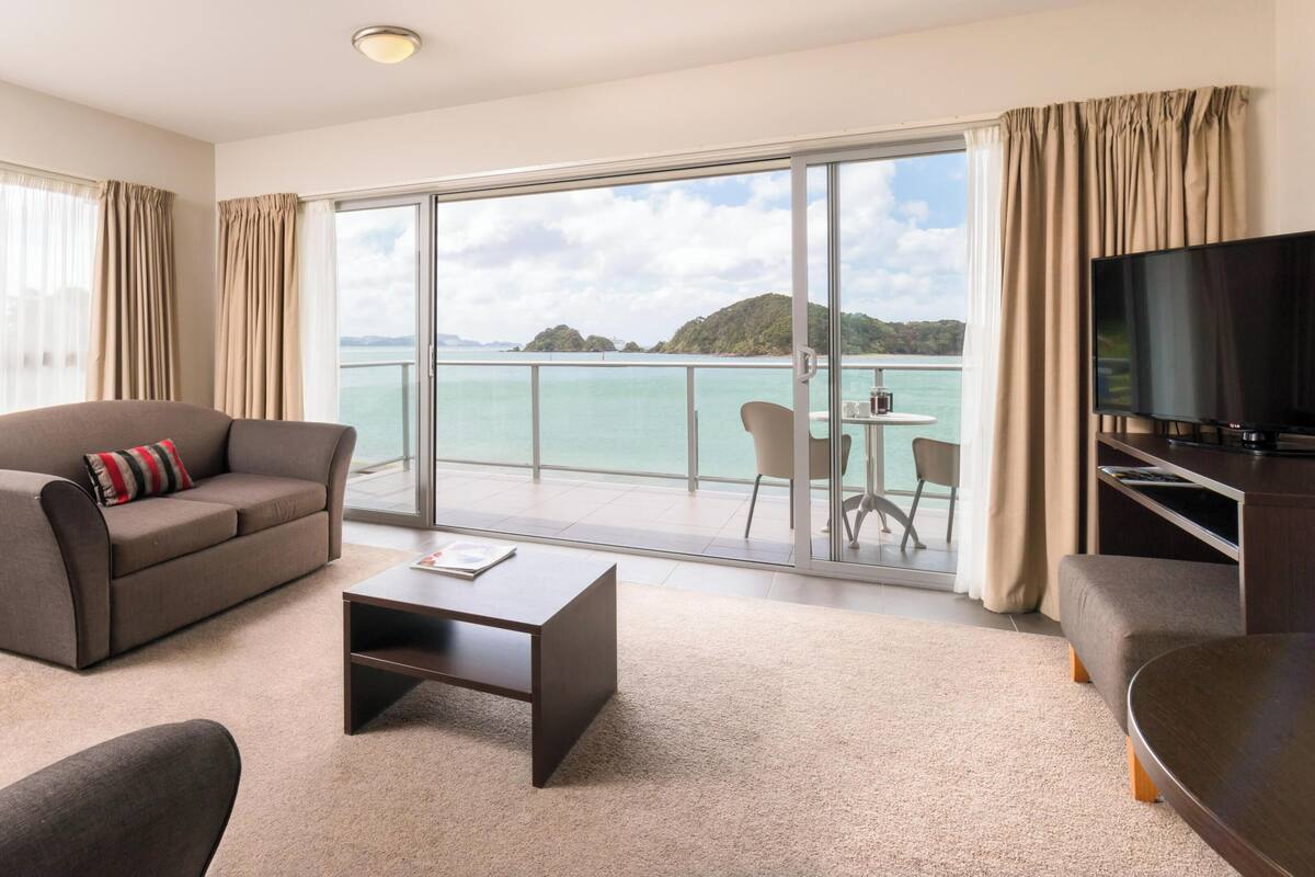 A modern hotel room with a sofa, a coffee table, a TV, and a balcony with a view of the sea and islands.