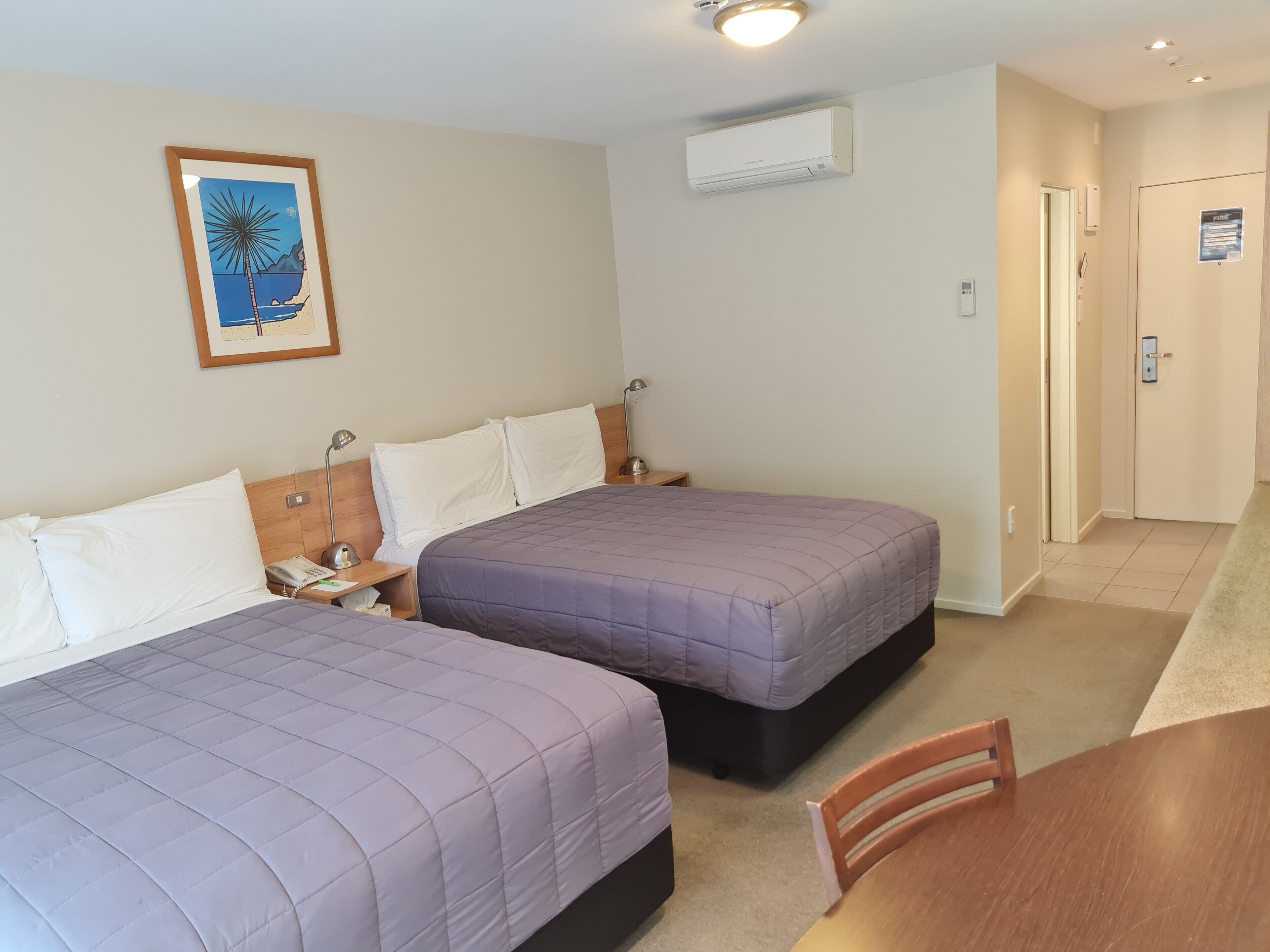 Ein Hotelzimmer mit zwei Betten, einem gerahmten Bild an der Wand, einer Klimaanlage und einer Tür mit einem Brandschutznachweis.