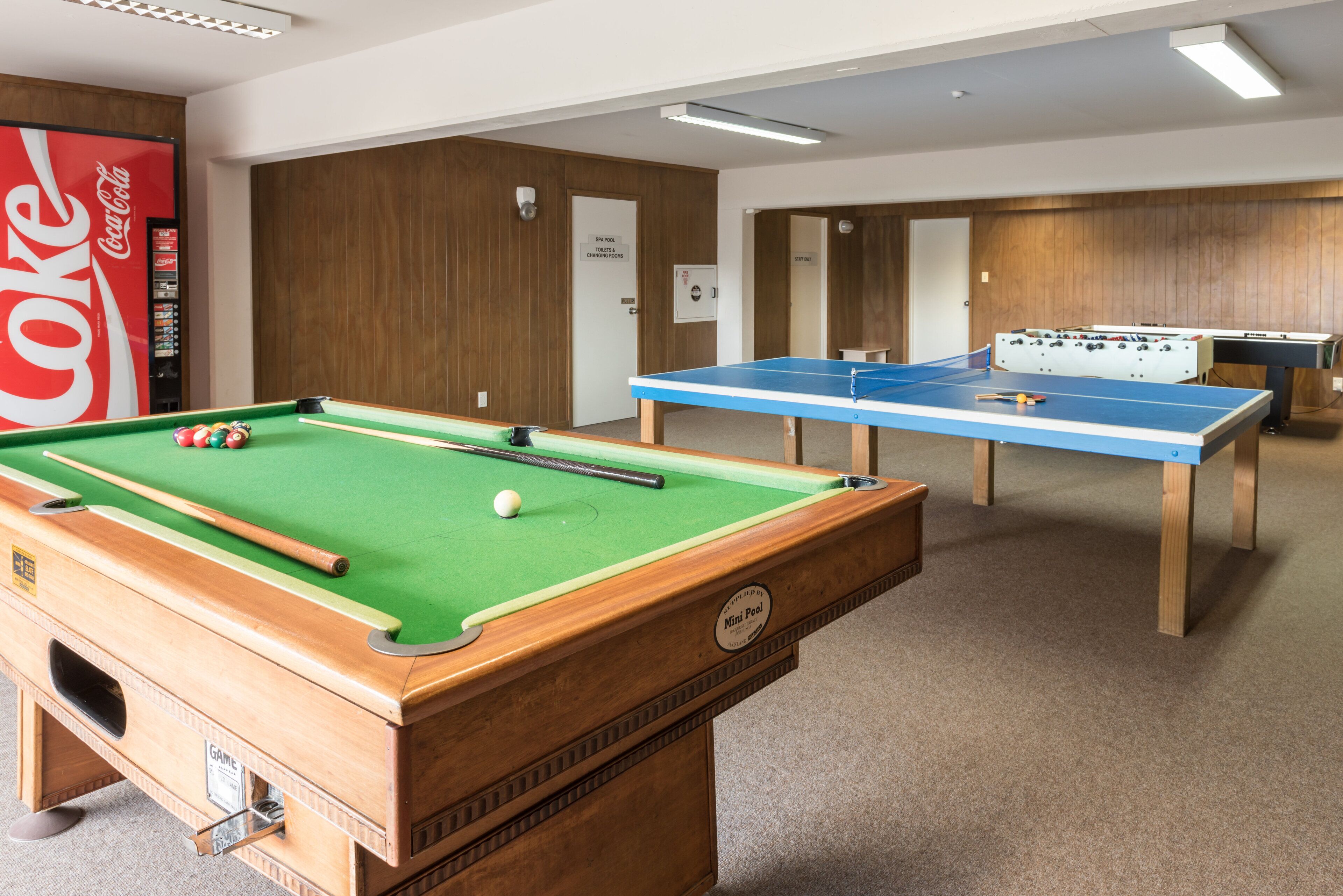 A pool table with billiard balls, a foosball table, and a Coca-Cola vending machine.