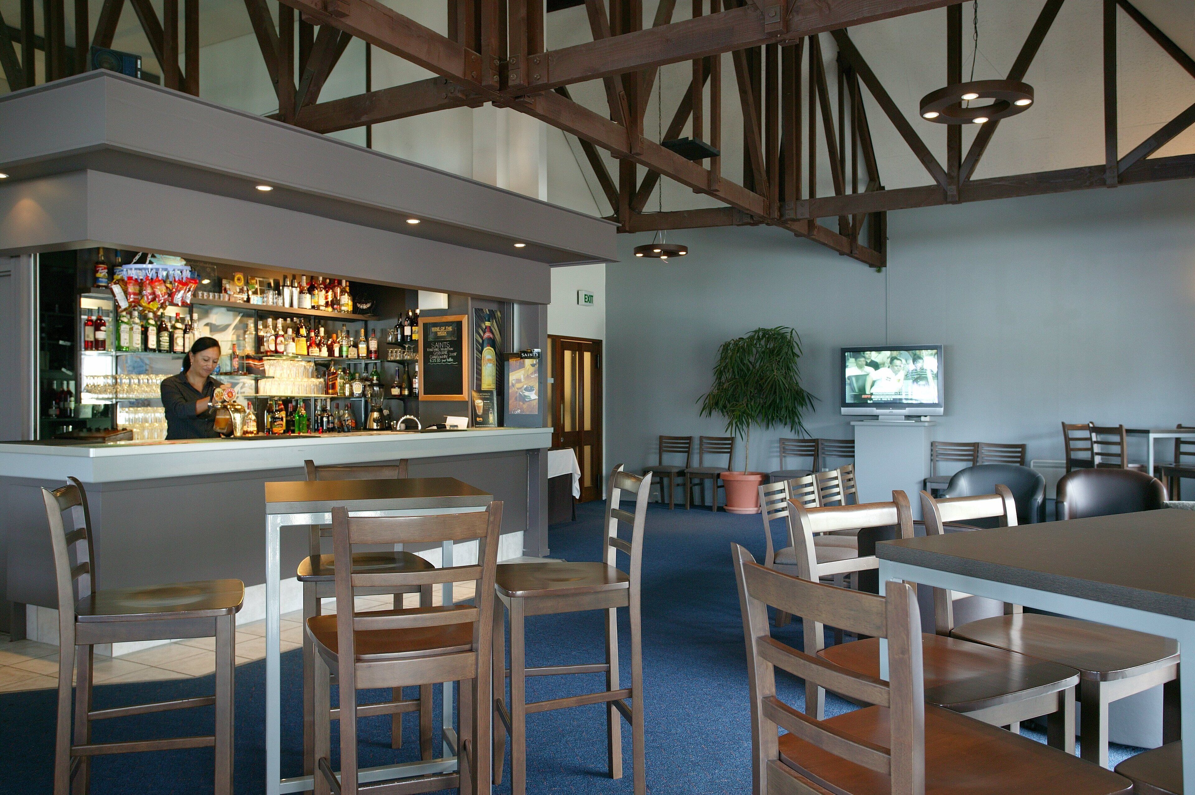 A bar area with wooden chairs and tables, a bar counter with various bottles, and a television mounted on the wall.