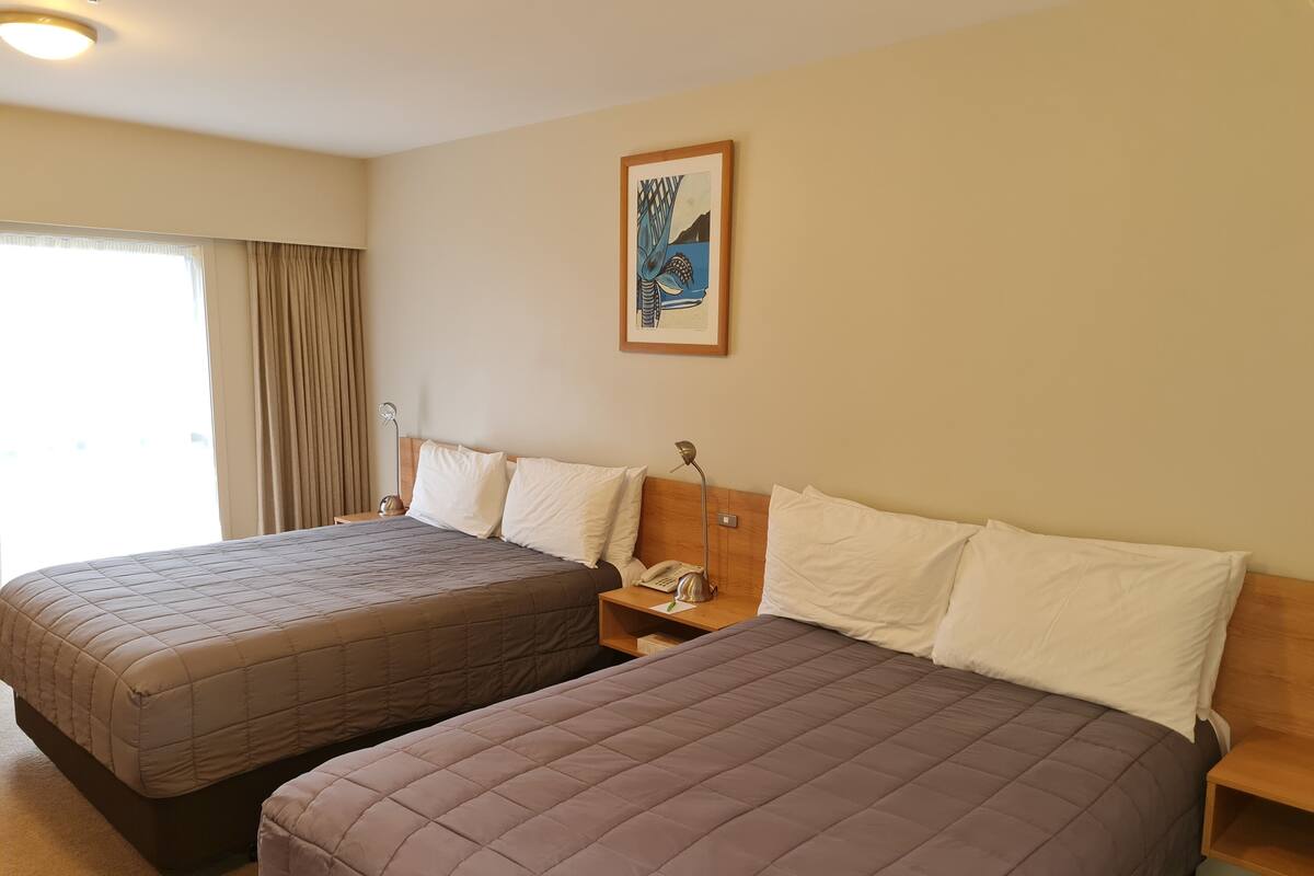 A hotel room with two beds, a nightstand with a lamp, a painting on the wall, and a window with curtains.