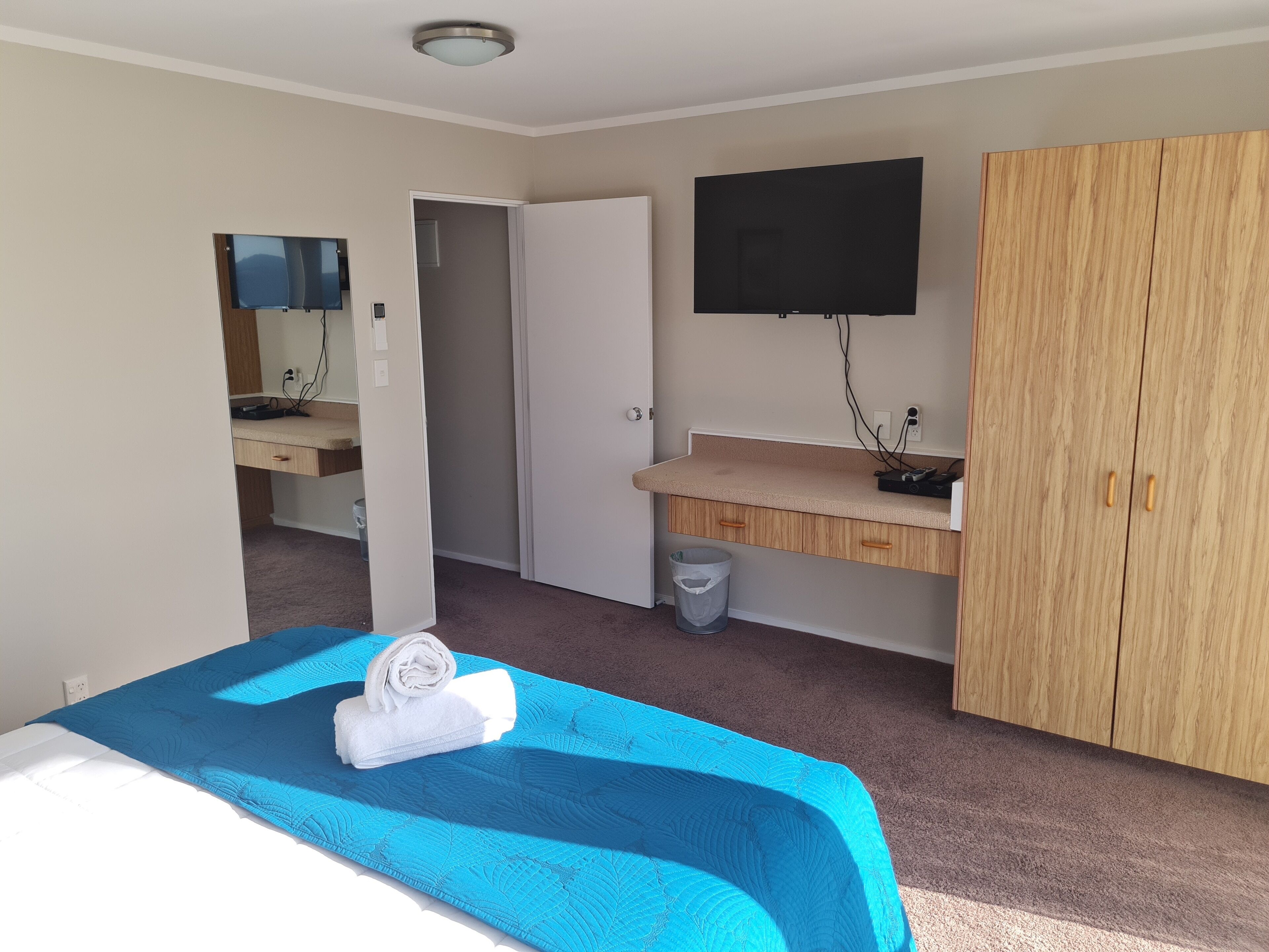 Habitación de hotel con cama, televisor montado en la pared, armario de madera, baño pequeño con espejo y lavamanos.