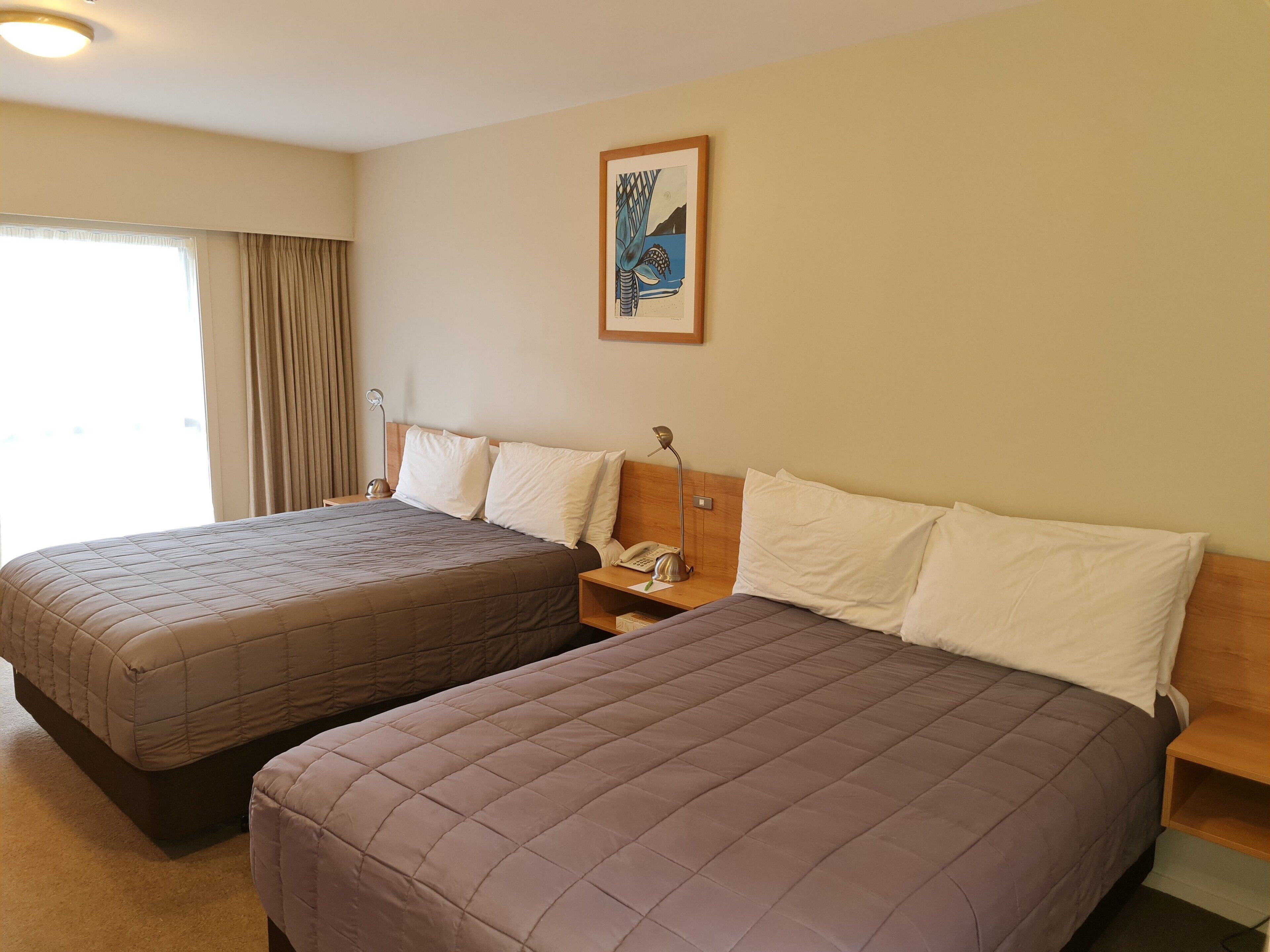 Una habitación de hotel con dos camas, una mesita de noche con lámpara, un cuadro enmarcado en la pared y una ventana con cortinas.