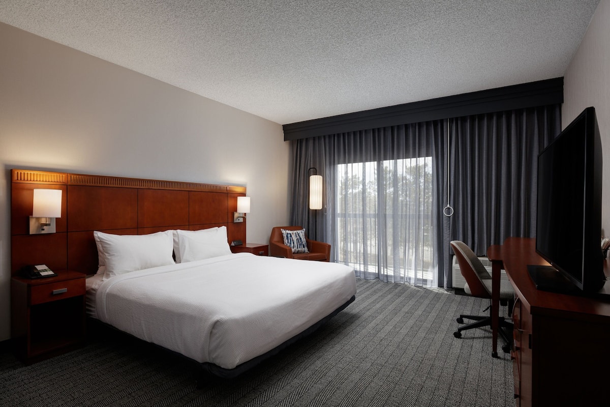 A hotel room with a large bed, a desk with a TV, a chair, a nightstand with a phone, and a window with curtains.