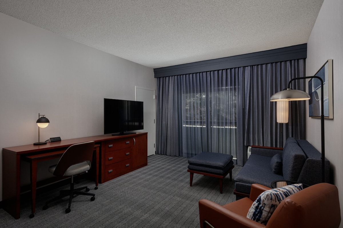 A hotel room with a wooden desk, a television, a sofa, a chair, a lamp, and a large window with curtains.
