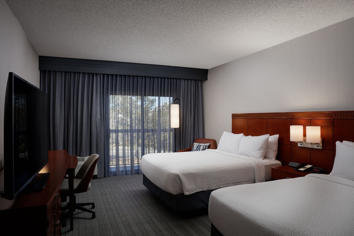 A hotel room with two beds, a desk with a chair, a TV, and a window with curtains.